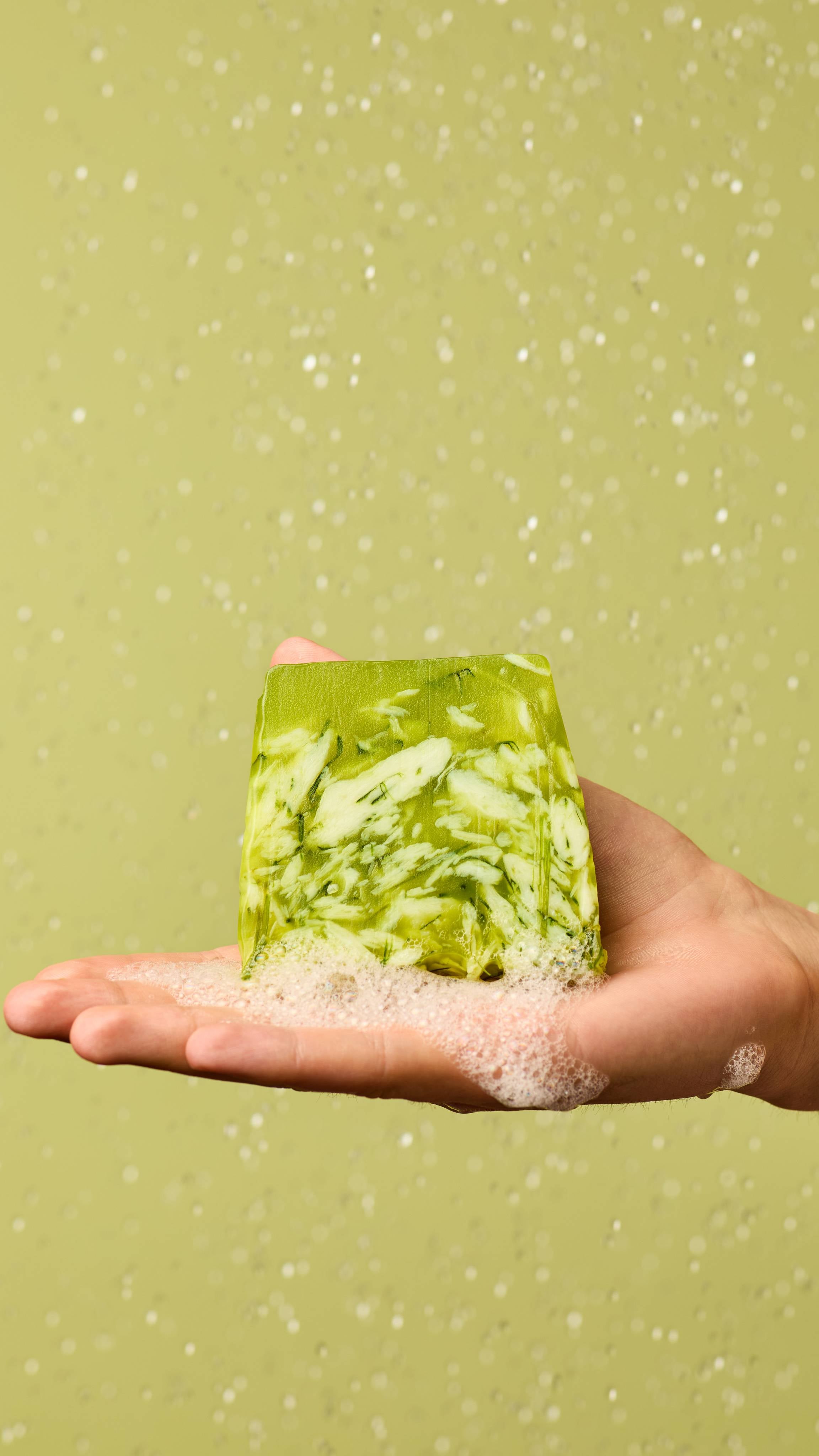 The image shows a light-toned hand holding an off-square slice of translucent green and white Lemongrass soap, marbled with inclusions. White foam gathers around the soap, with water droplets on a light green background.