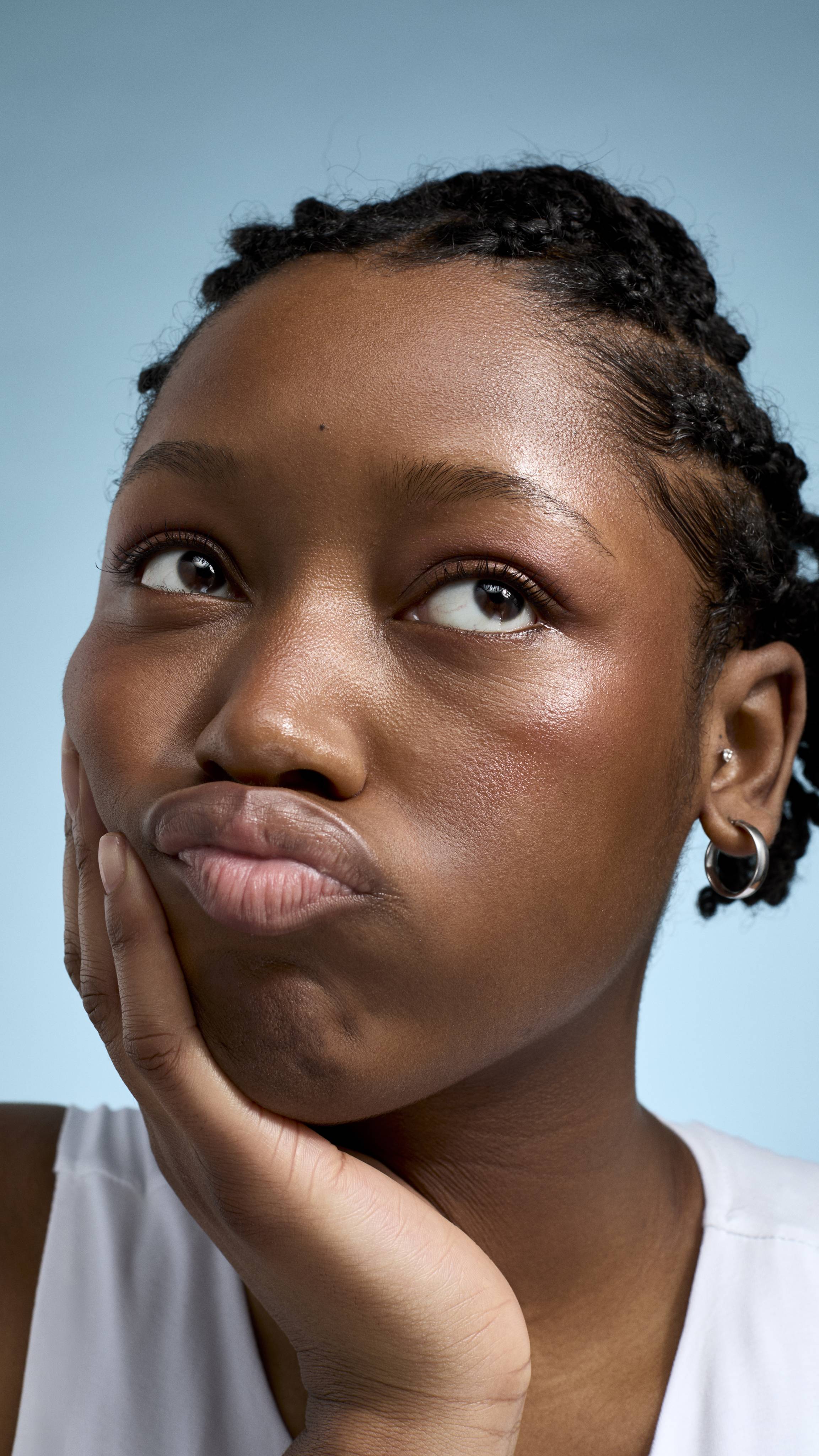 A close-up of a model with deep-toned skin and braided hair, looking upwards with a thoughtful, pouting expression. They are resting their chin in their hand, wearing a white top and small silver hoop earrings against a soft blue background.