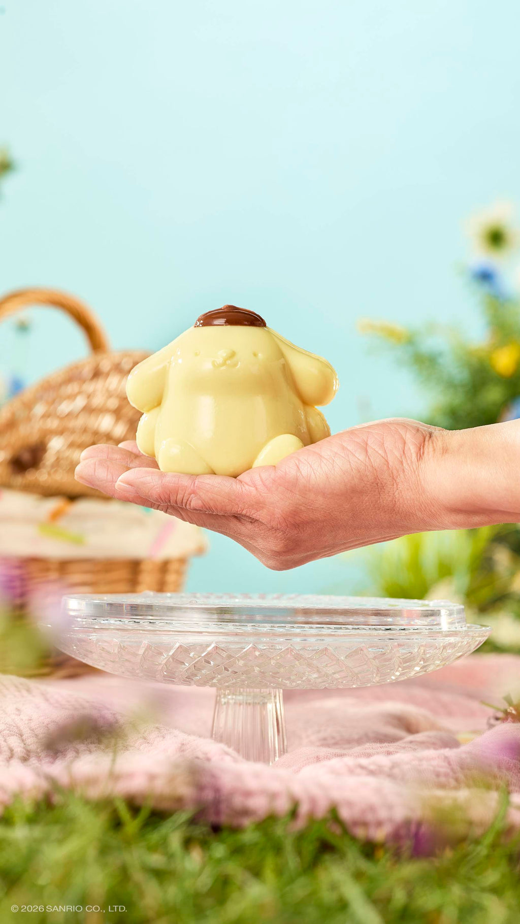 A hand holds a glossy, pale yellow "Pompompurin" shower jelly over a glass cake stand in a sunny meadow. The dog-like character wears a brown beret. A wicker basket and pink blanket sit on the grass under a blue sky.