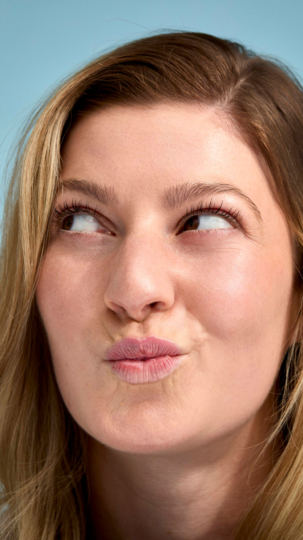 A close-up of a model with fair skin and dark blonde hair looking upwards with a playful, pouting expression. Their face has a natural, glowing finish, set against a soft, light-blue background.