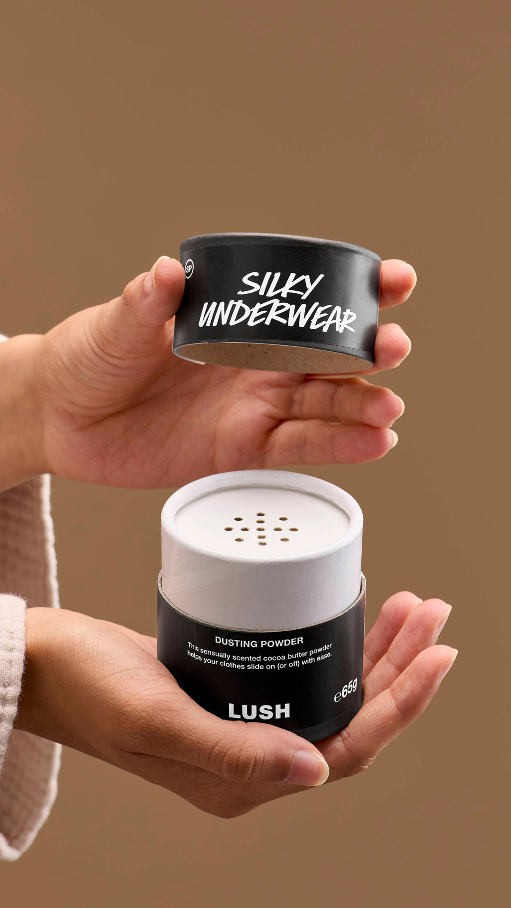 The image shows a pair of hands opening a black and white cardboard tub of 'Silky Underwear' dusting powder. The tub has perforated holes on top, with a black lid with white text, against a solid beige background.