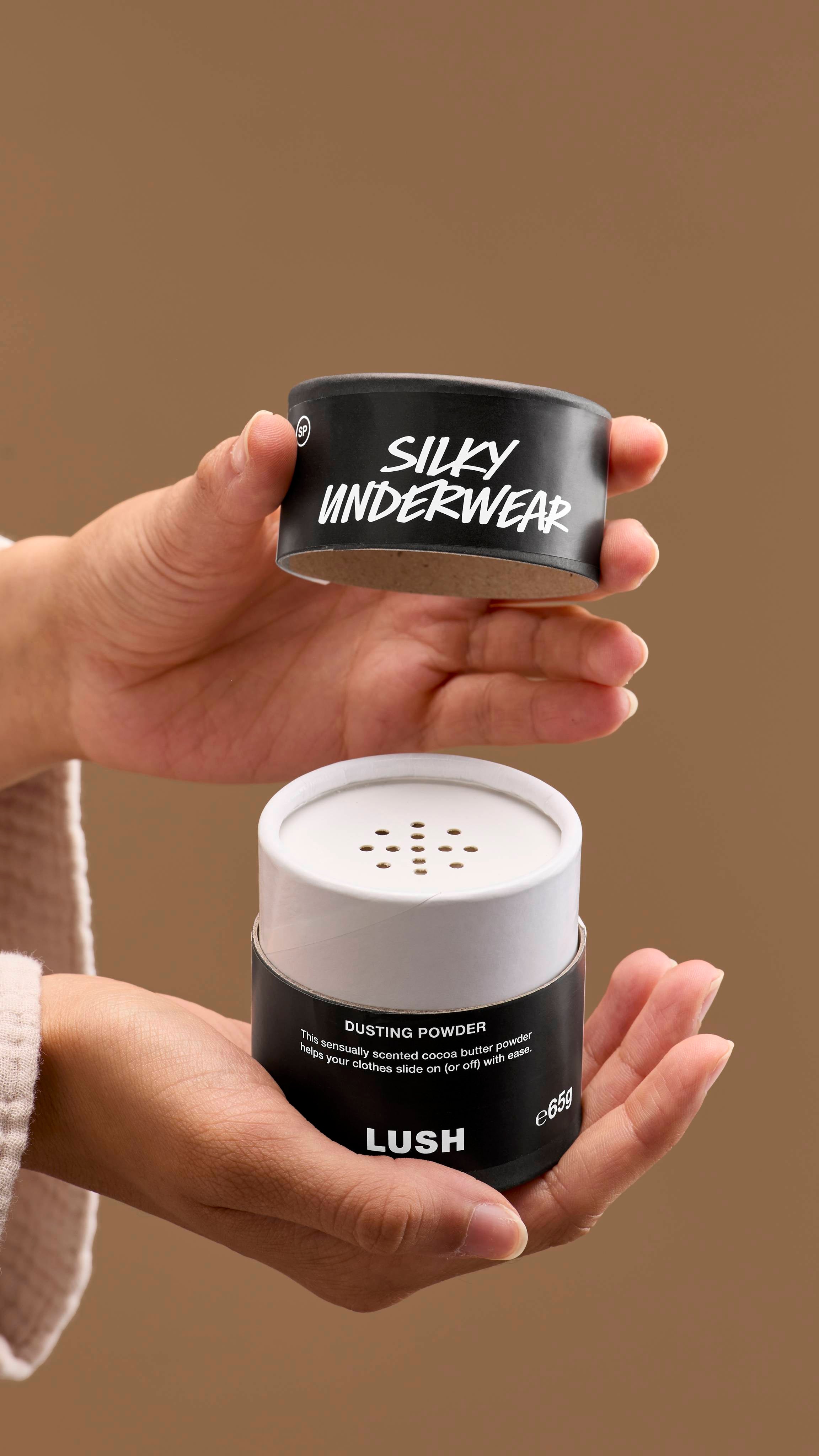The image shows a pair of hands opening a black and white cardboard tub of 'Silky Underwear' dusting powder. The tub has perforated holes on top, with a black lid with white text, against a solid beige background.