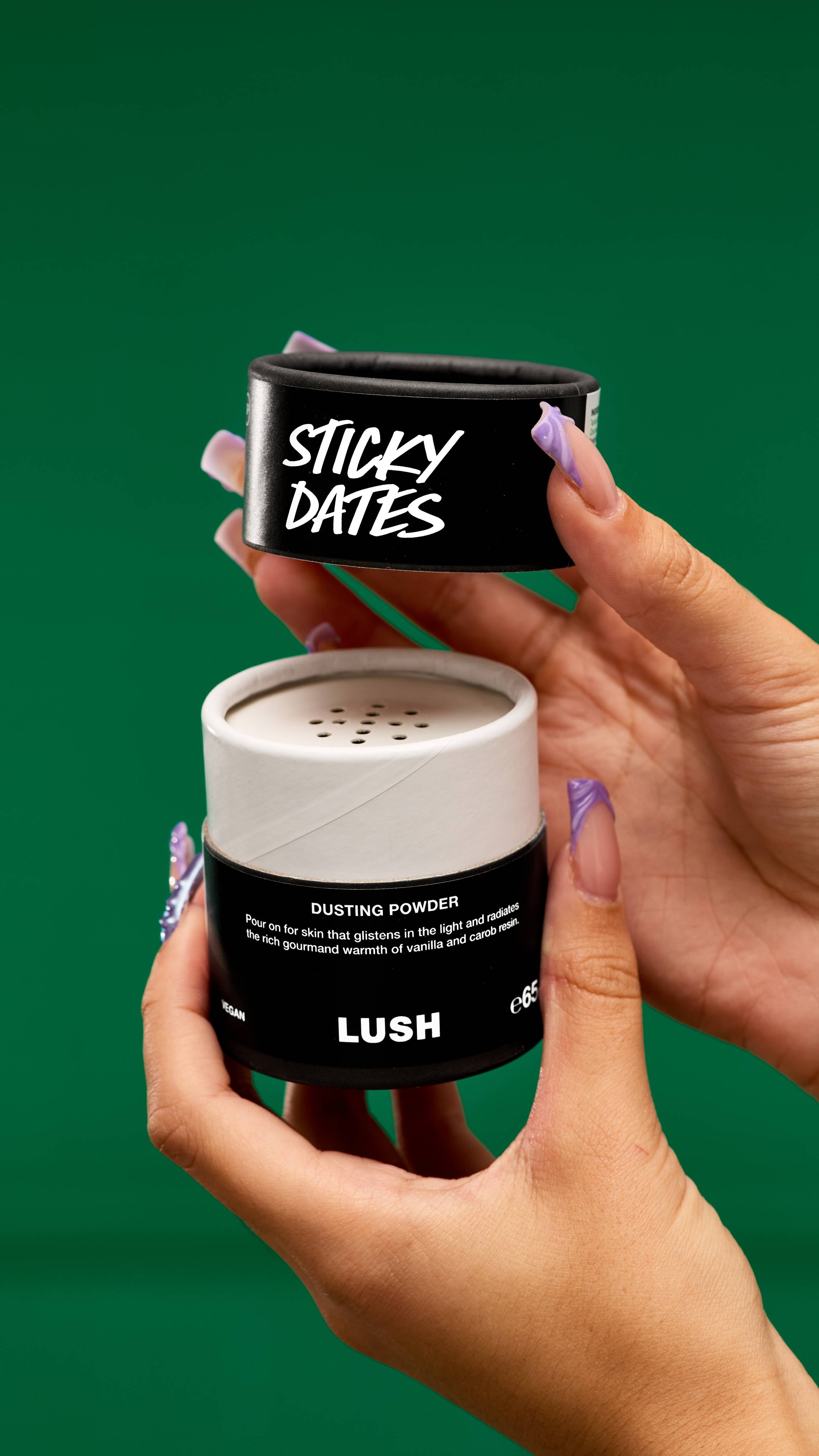 A pair of hands holds open a LUSH Sticky Dates dusting powder container with a black lid and white label. The white powder inside is visible against a vibrant green background.