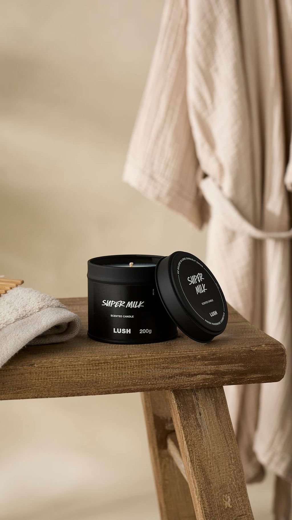 A slightly off-center shot of the black Super Milk Scented Candle tin, sitting open on a rustic, weathered wooden stool. The lid rests leaning on the tin, and a light-coloured towel and a beige bathrobe hang in the soft-focus background.