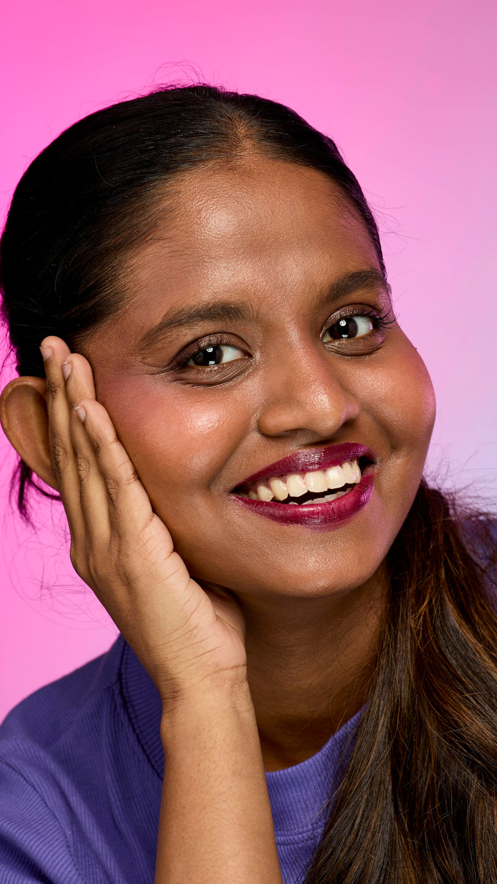 A smiling person with dark hair and skin, wearing a purple top, is shown from the chest up. They are wearing the Lush, deep plum Tartlet lip cream and have one hand touching the side of their face. The background is a pink and purple gradient.