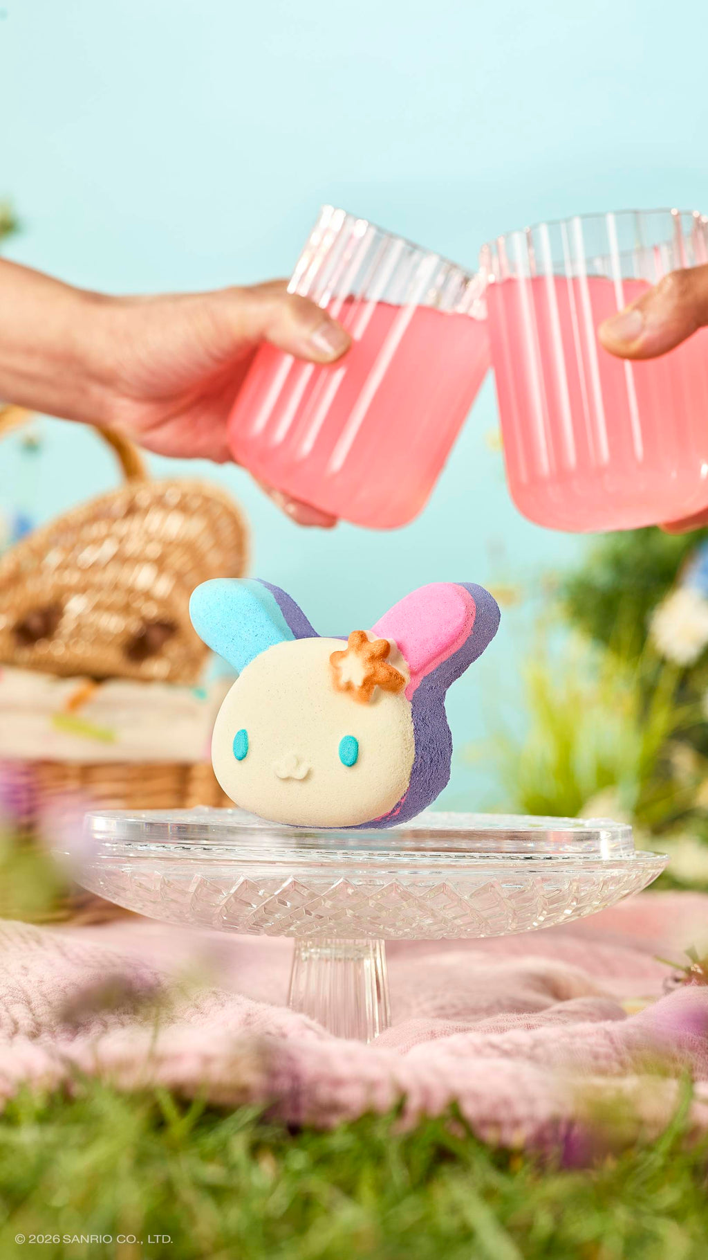 The bunny-shaped "U.SA.HA.NA" bath bomb with blue and pink ears sits on a glass cake stand in a meadow. Above it, two hands clink glasses of pink juice in a toast. The sunny picnic scene includes a wicker basket and a soft blue sky.