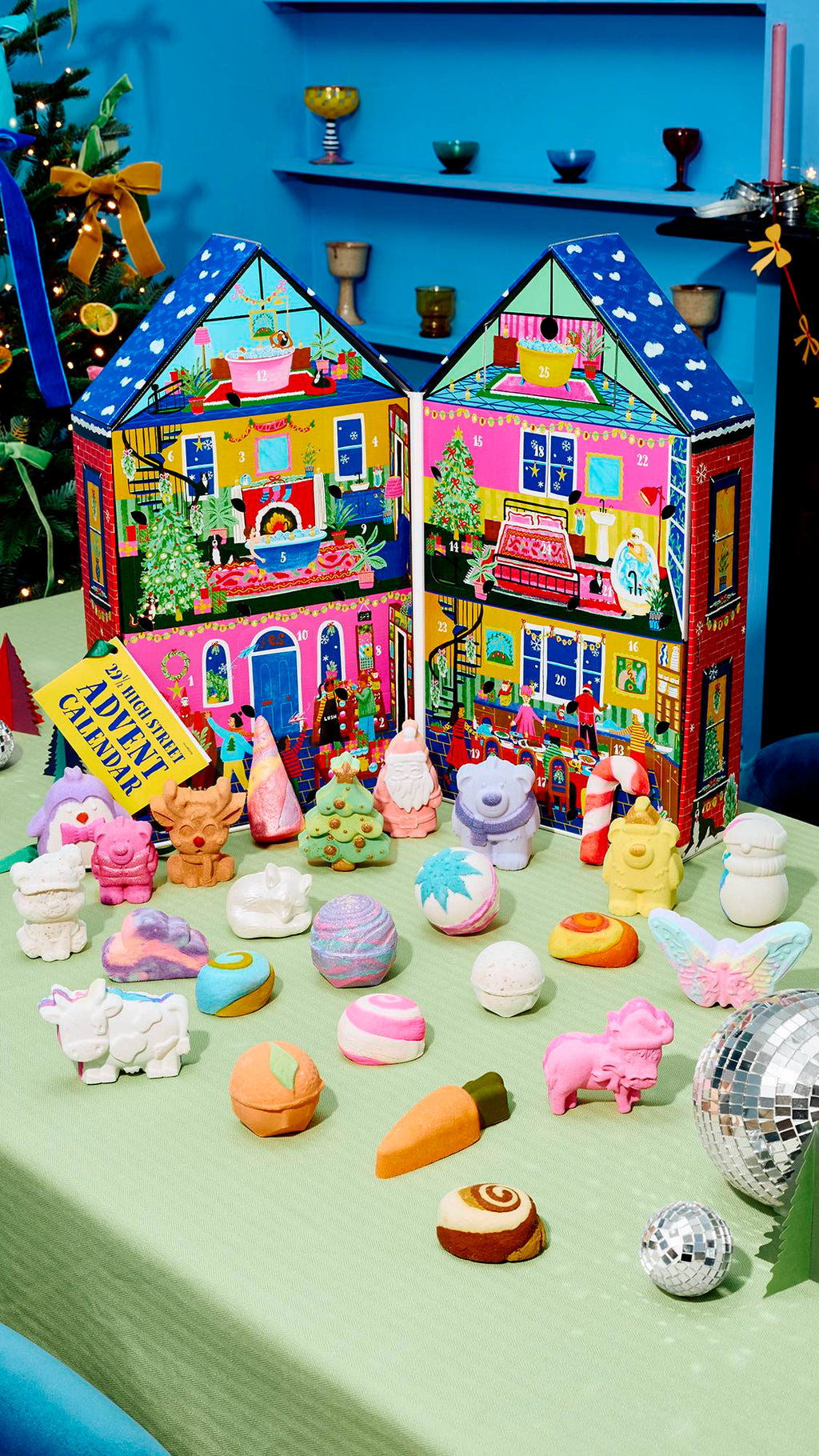 An unfolded house-shaped advent calendar sits open on a light green table, surrounded by an array of colourful festive bath bombs and soaps, including a reindeer, Santa, and a polar bear, with disco balls catching light in the foreground.
