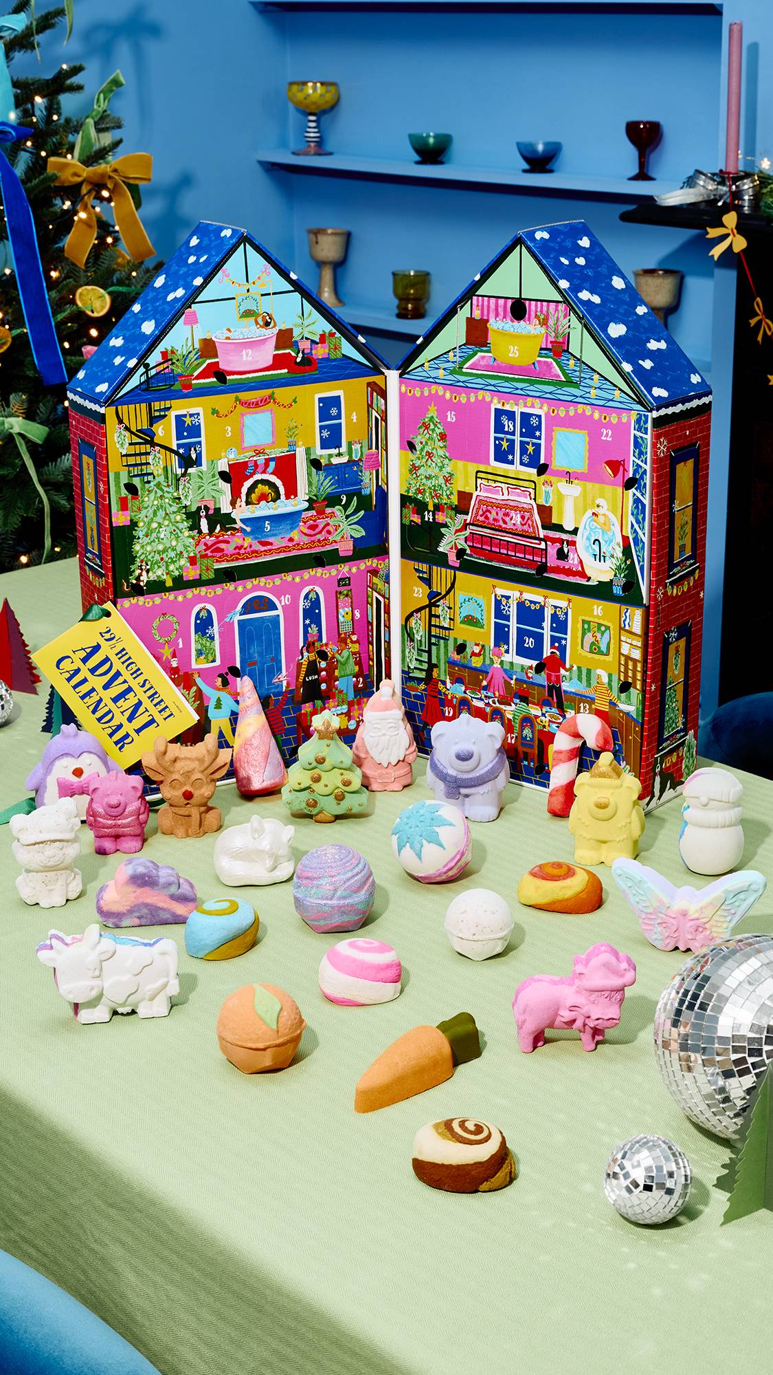 An unfolded house-shaped advent calendar sits open on a light green table, surrounded by an array of colourful festive bath bombs and soaps, including a reindeer, Santa, and a polar bear, with disco balls catching light in the foreground.