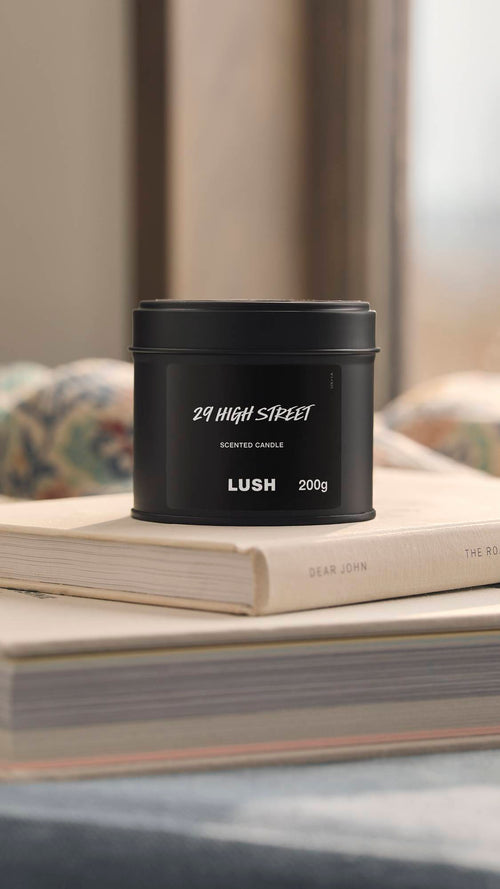 The image shows a natural, bright bedroom background. The foreground shows the black tin 29 High Street candle sitting on top of a small pile of books.