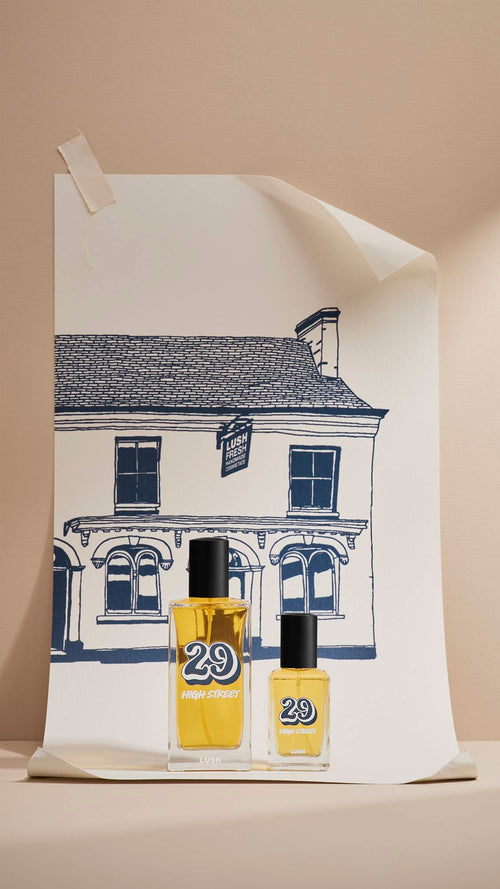 Two glass bottles of Lush '29 High Street' perfume, 30ml and 100ml, stand in front of a large cream poster. The poster features a navy-blue line drawing of the original Lush shop. The scene is set against a neutral beige background.