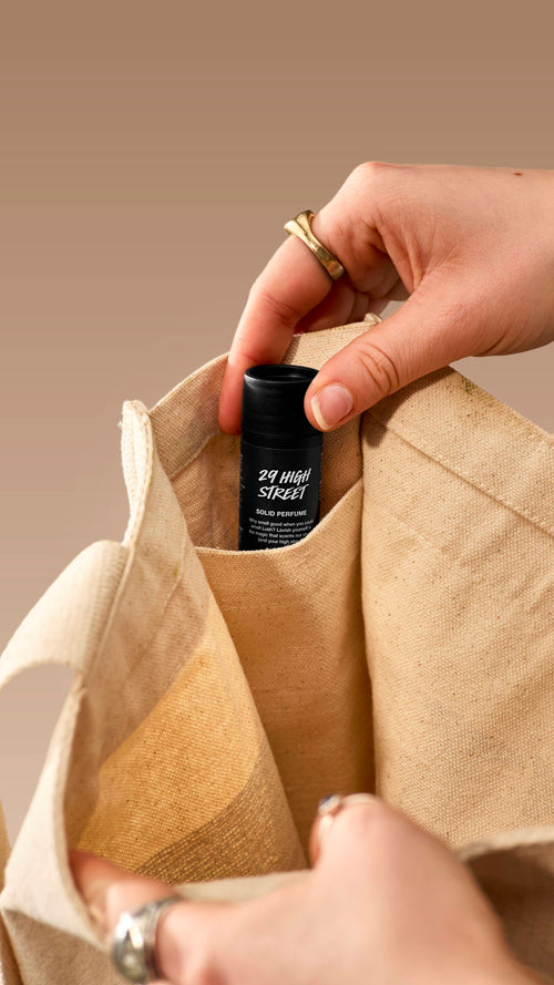 A close-up shot shows a hand placing a small, black Lush '29 High Street' solid perfume tube into the inner pocket of a cream canvas tote bag. The person is wearing several rings, and the image is set against a neutral beige background.
