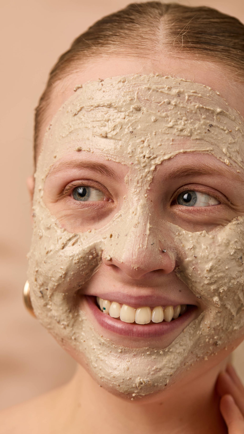 A close-up image of the model's face on a warm, beige background. Their copper hair is tied back, and they have applied the textured Bring Us Some Figgy Pudding face mask all over their face as they smile.