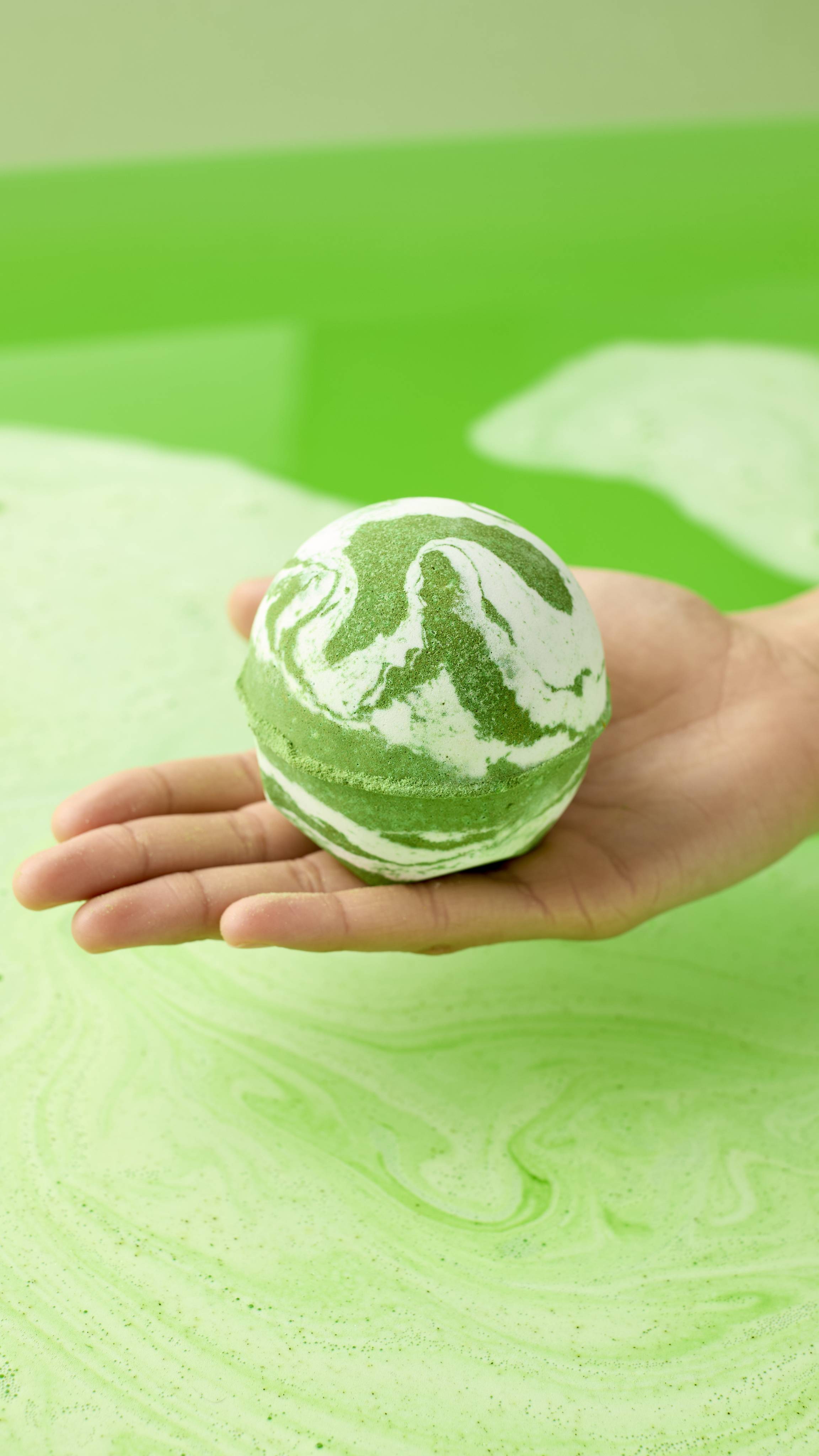 A hand holds the green and white marbled Cold Bath Bomb over a bathtub filled with vibrant green water. The bathwater swirls with light green foam, perfectly matching the colors of the bath bomb.