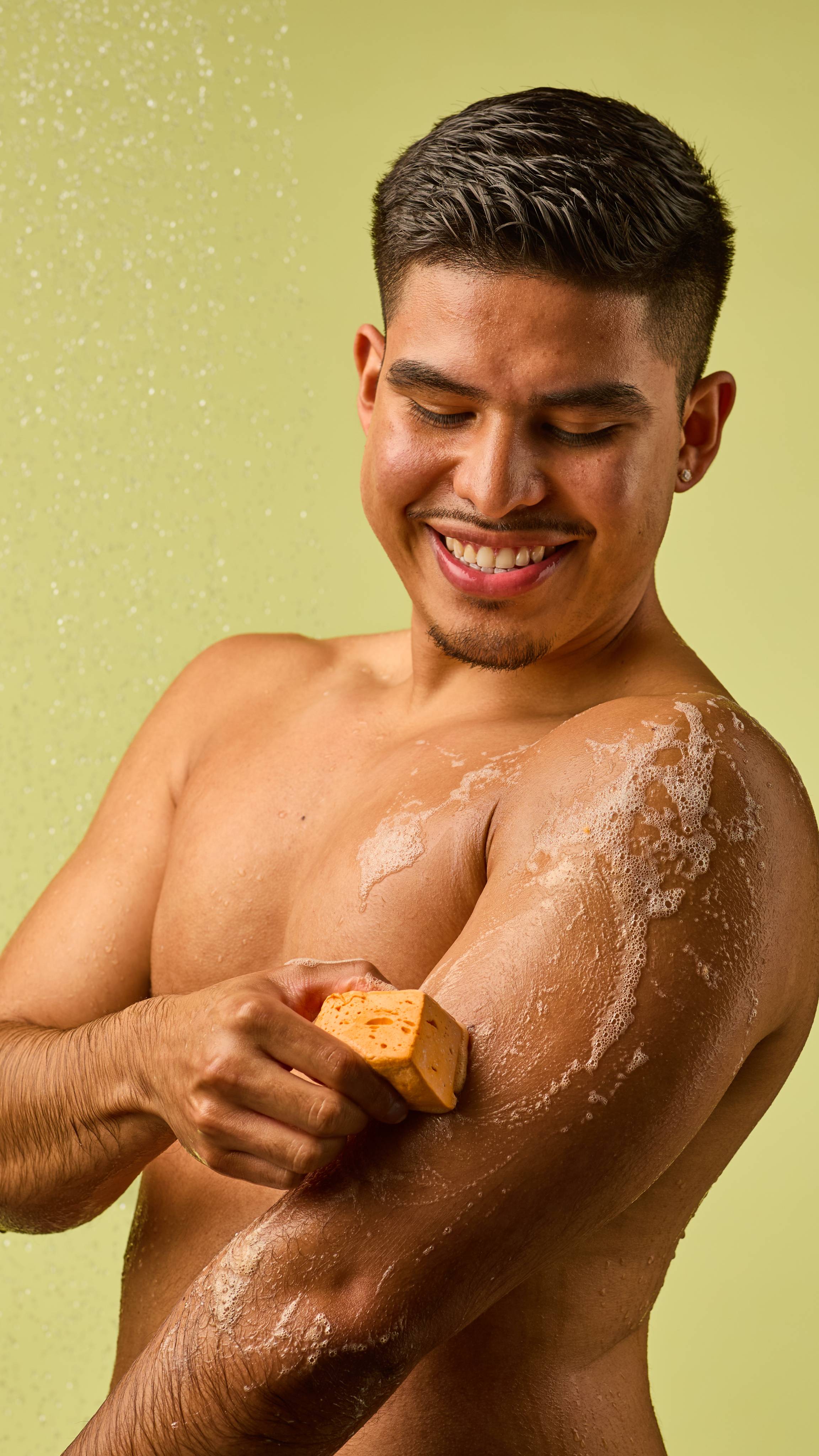 The image shows a person with short dark hair, smiling. They rub the pastel orange Passion Fruit Delight floating soap onto their arm, creating white, bubbly lather. Water droplets fall on them against a light green background.