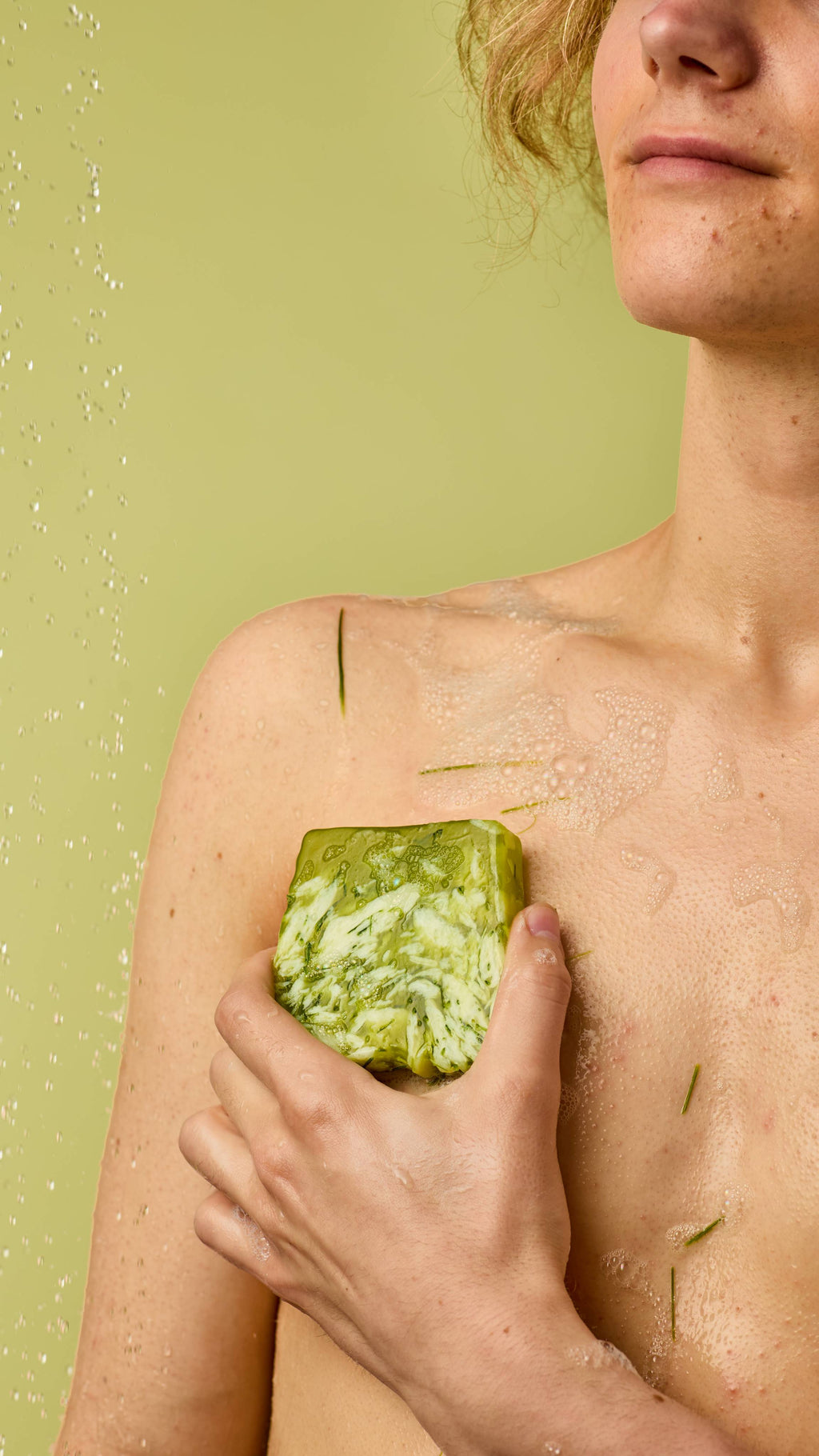 The image shows a light-skinned model standing on a pastel-green background with water droplets falling. The model is using the green, marbled Lemongrass soap over their torso and shoulder to create a soft lather of foam.