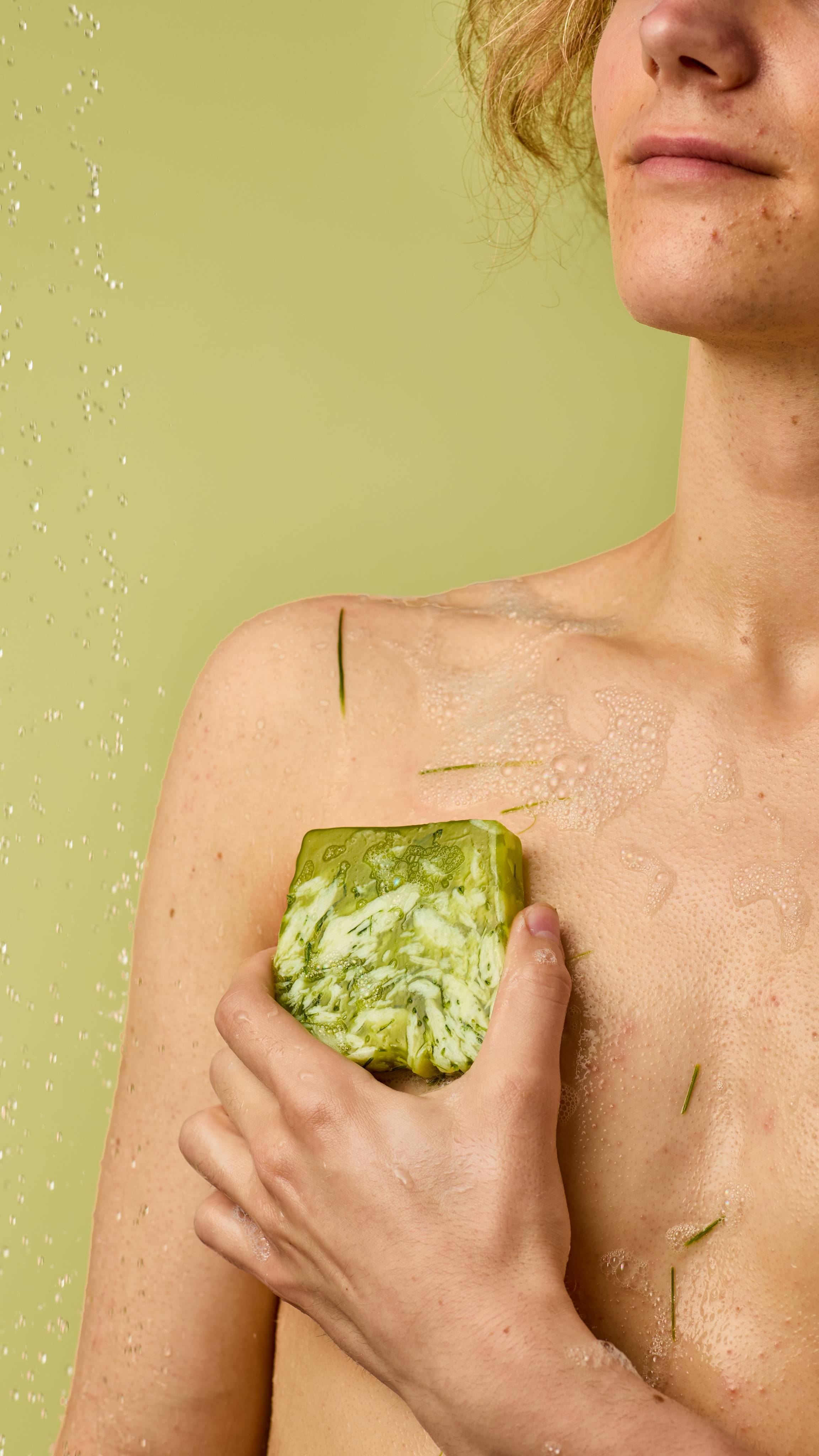 The image shows a light-skinned model standing on a pastel-green background with water droplets falling. The model is using the green, marbled Lemongrass soap over their torso and shoulder to create a soft lather of foam.