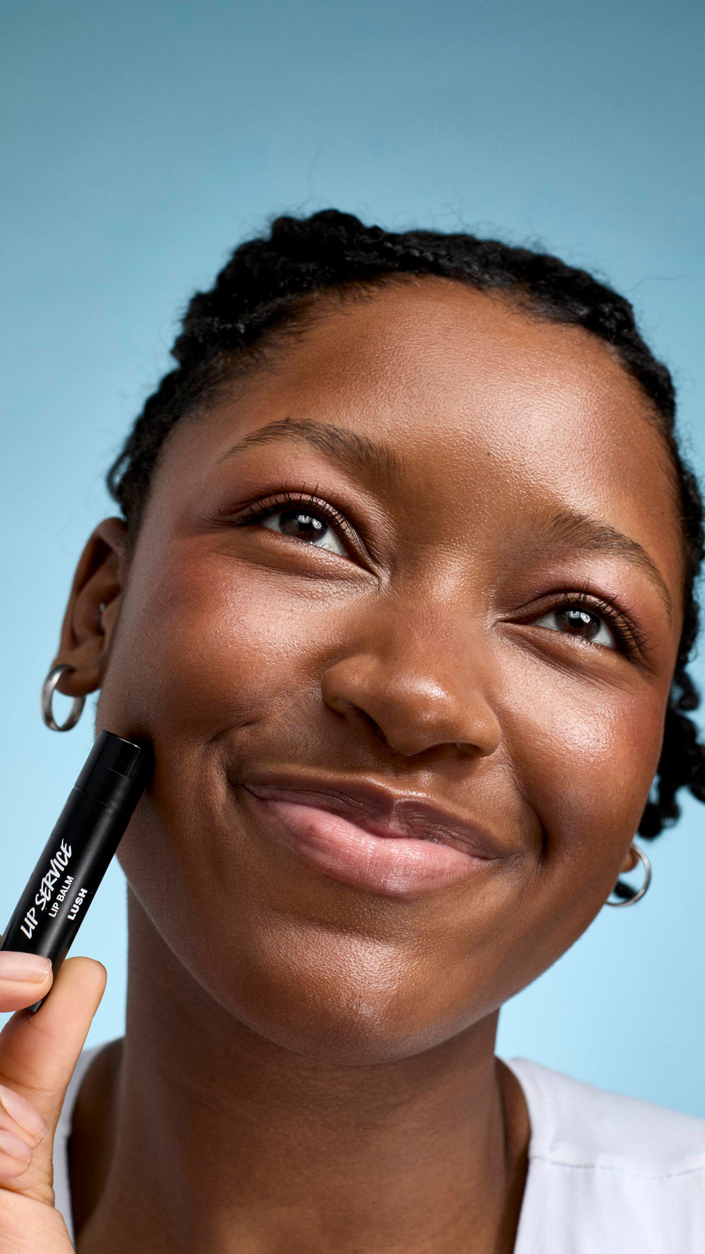 A close-up of a model with braided hair and glowing skin smiling warmly. They hold a black tube of Lush 'Lip Service' lip balm against their cheek while looking slightly off-camera against a soft blue background.