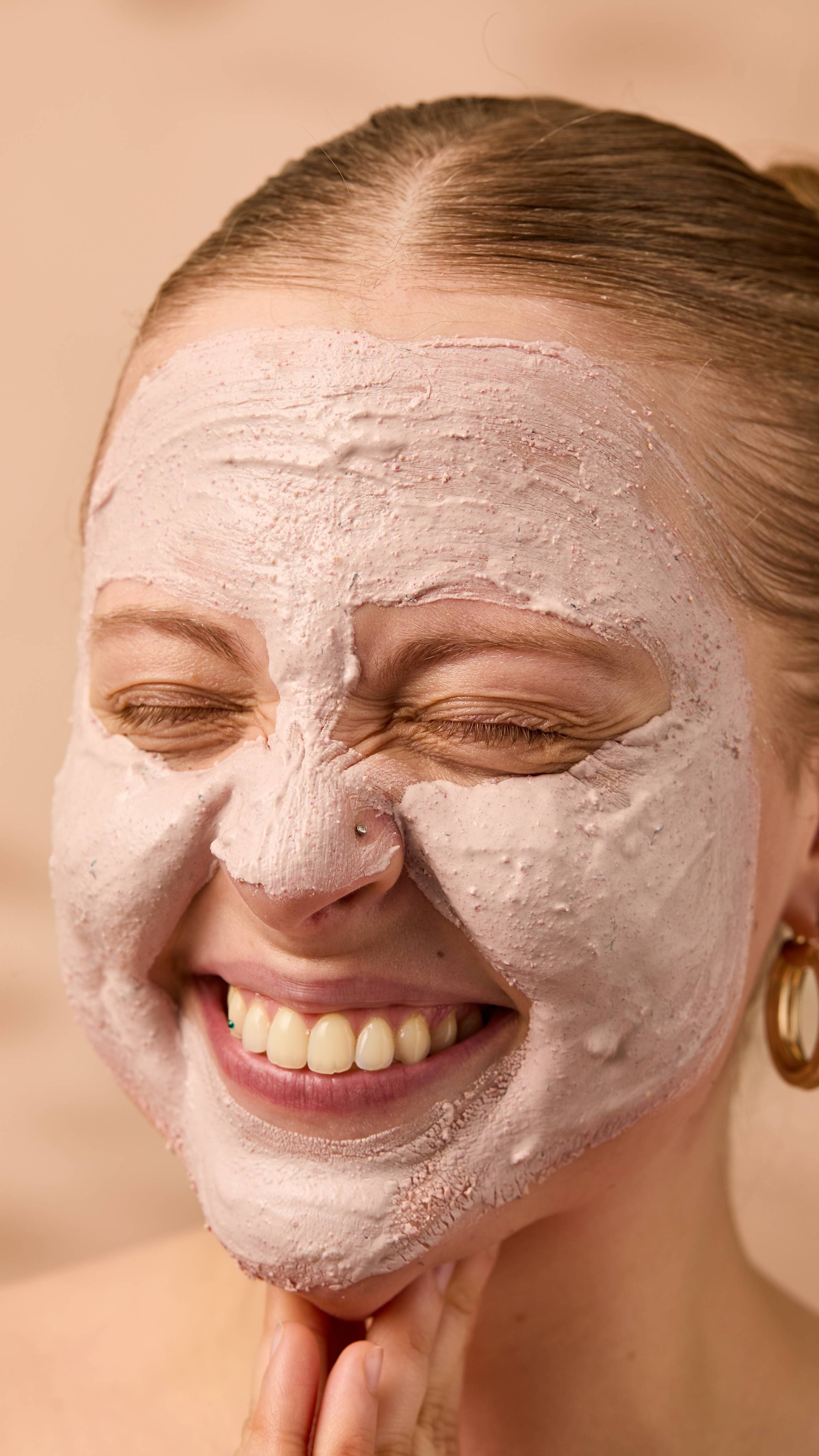 A close-up image of the model's face on a warm, beige background. Their copper hair is tied back, and they have applied the thick Mrs Whippy face mask over their entire face as their eyes are closed and they smile.