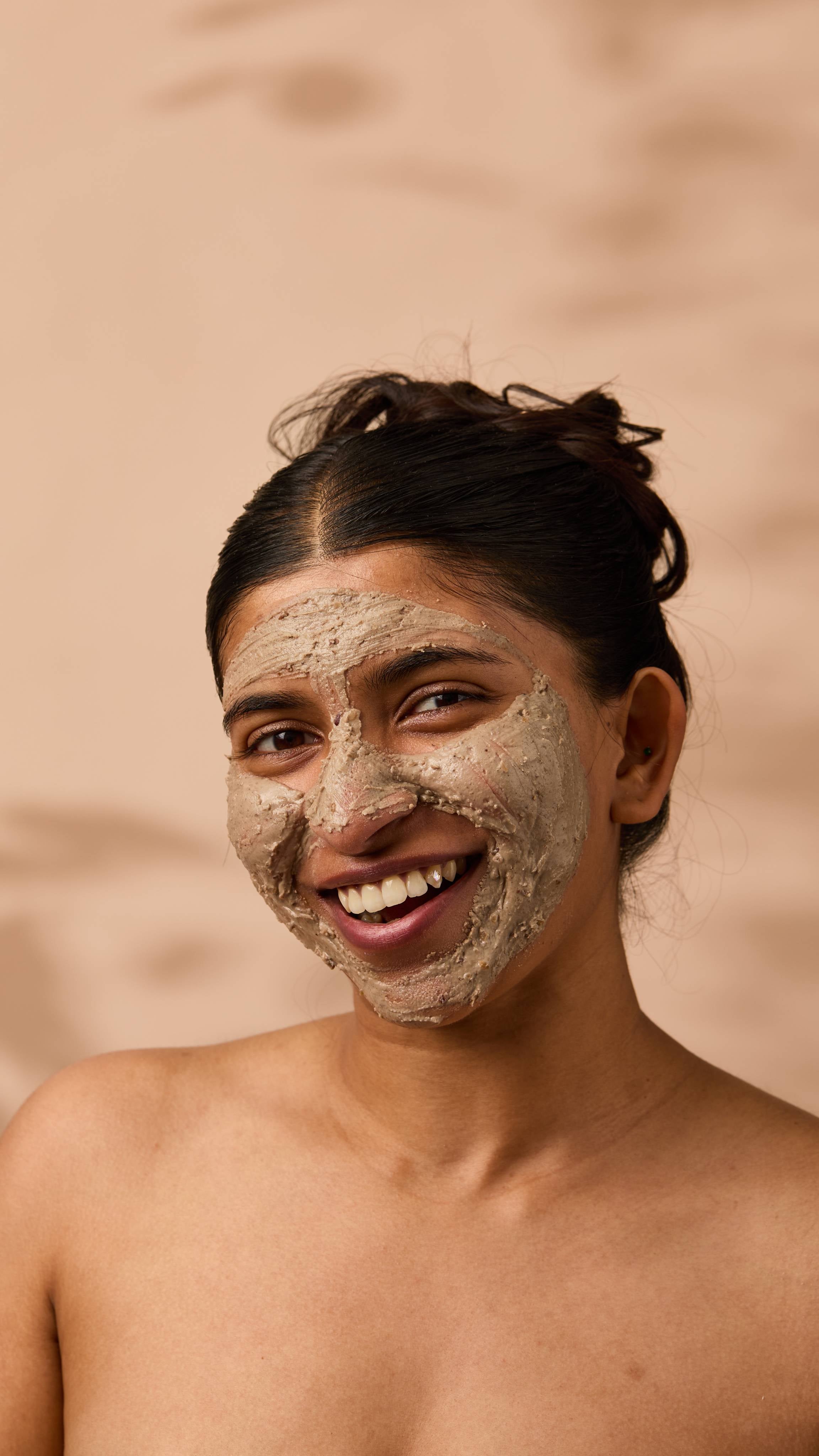 An image of the model's face on a warm, beige background. They have a smudge of the thick, creamy Nutkins face mask over their entire face as they smile.