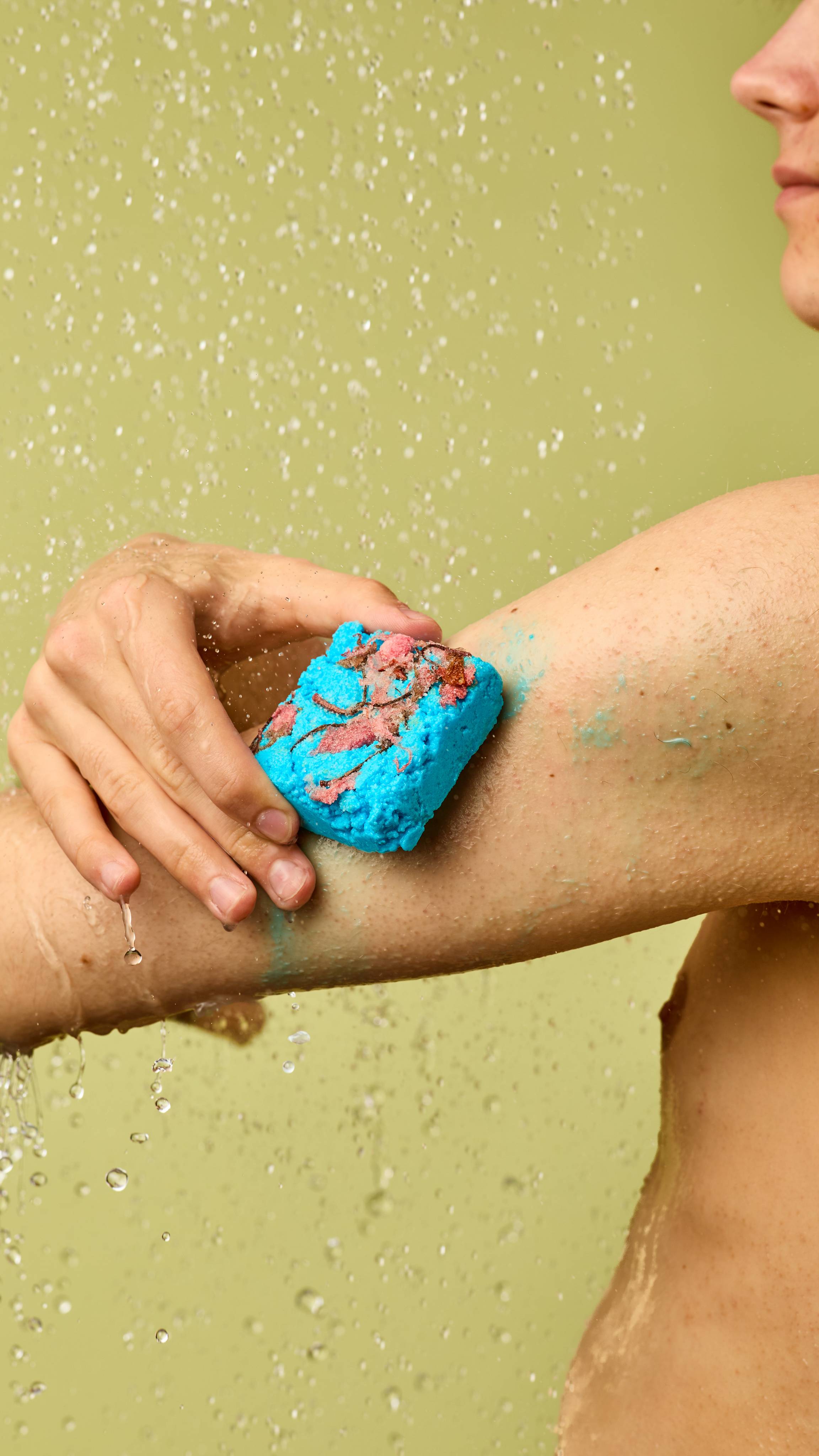 The image shows a person rubbing the square, vibrant turquoise Rub Rub Rub solid body scrub, with a rough brown patch and salt crystals, onto their arm. Light blue residue and water droplets are on their skin against a light green background.