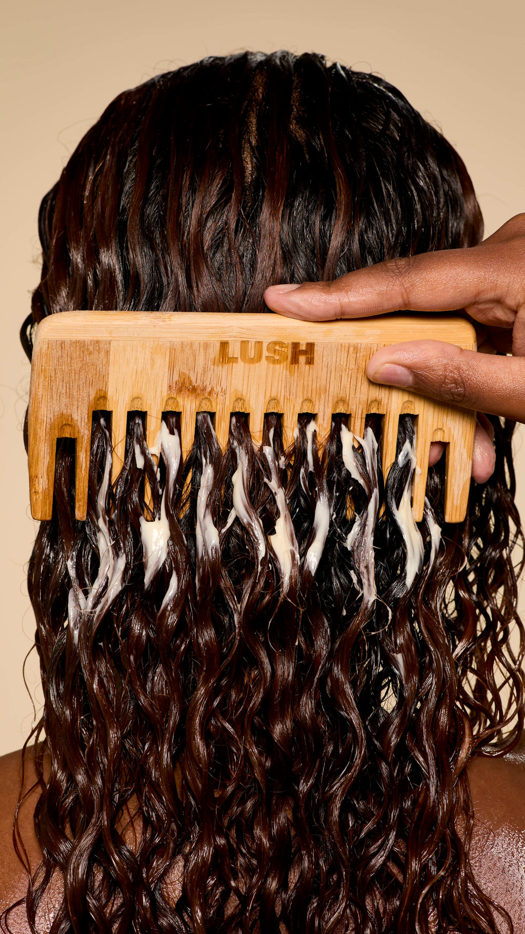 From behind, a hand uses a wooden Lush comb to detangle wet, dark, curly hair generously coated with a creamy Super Milk conditioner, creating beautiful, defined ringlets.