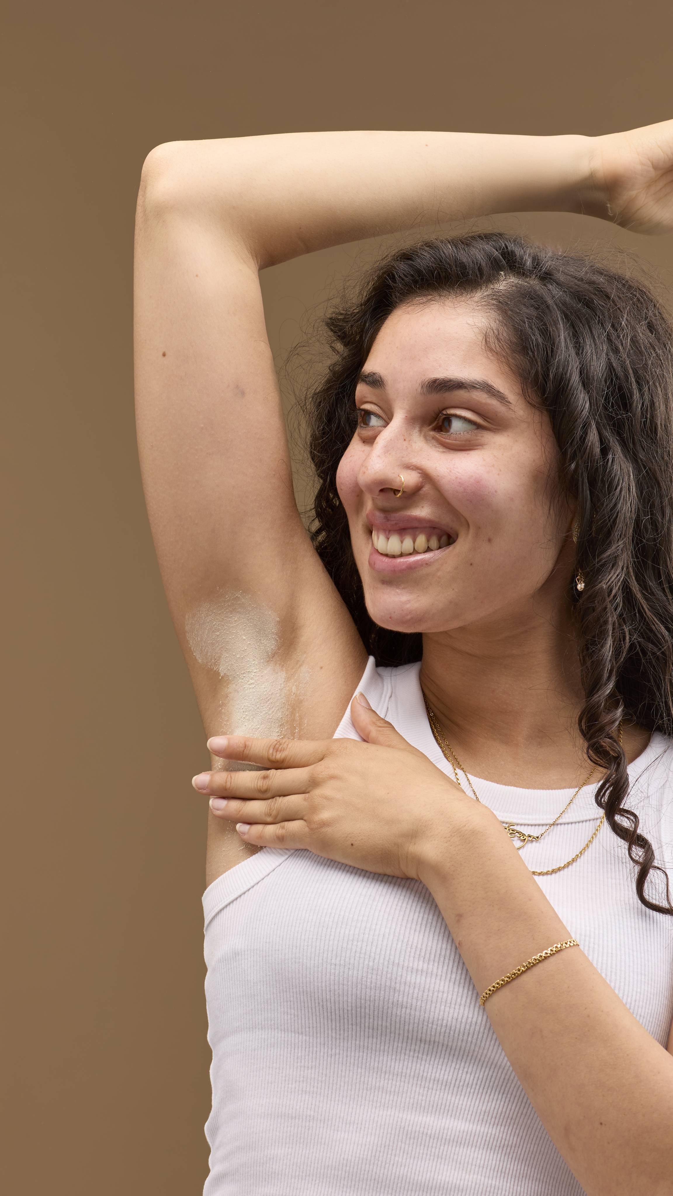 The image shows a person with dark, curly hair, smiling, with a raised arm. A pale cream 'The Greeench' deodorant powder is dusted onto the underarm. The person wears a white vest top and necklaces, against a light beige background.