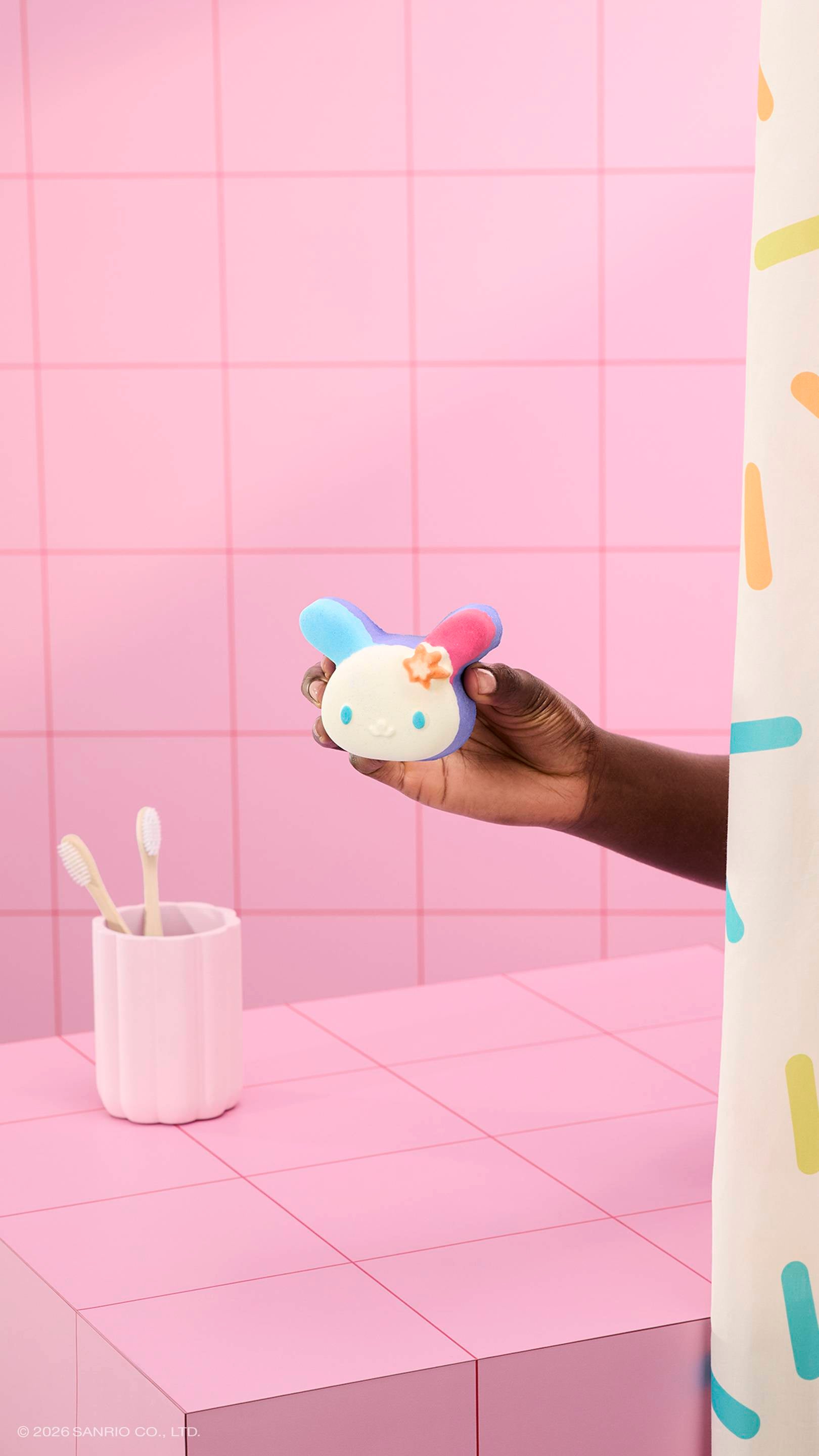 A hand holds a colourful bunny-shaped "U.SA.HA.NA" bath bomb in a pink tiled bathroom. A pink cup with two toothbrushes sits on the counter, and a sprinkle-patterned shower curtain is visible on the right.