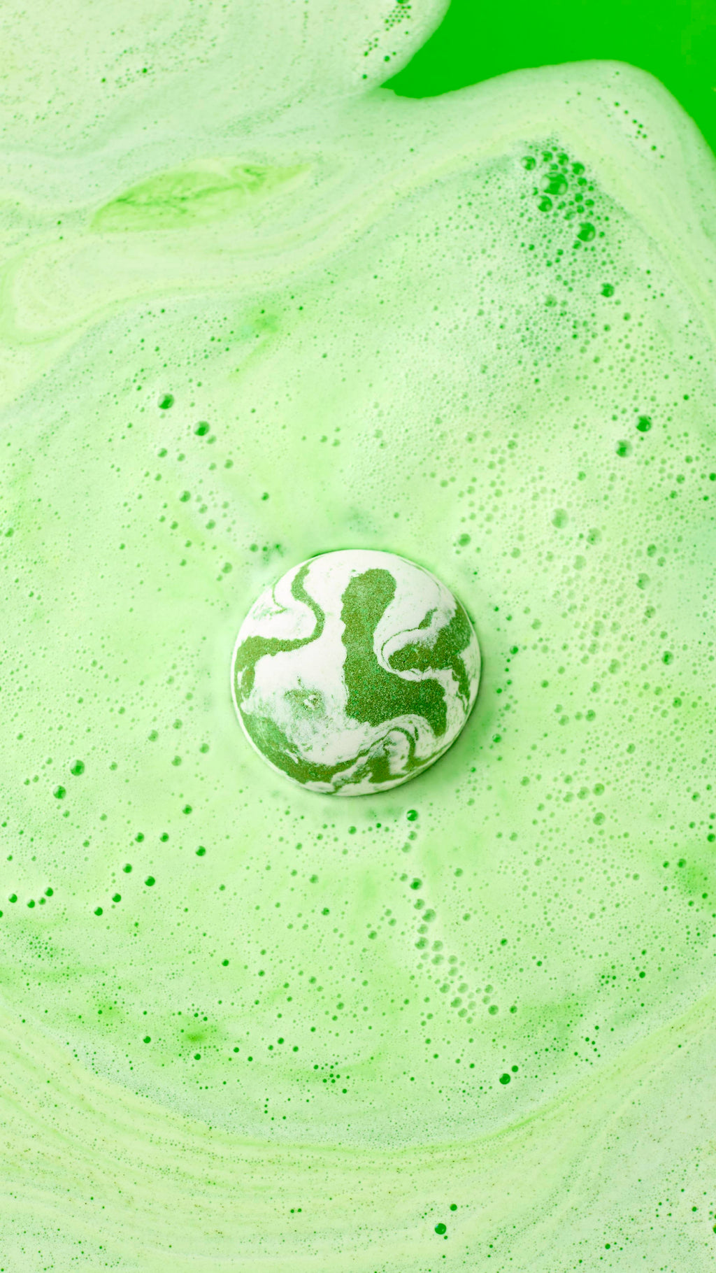 A hand holds the green and white marbled Cold Bath Bomb over a bathtub filled with vibrant green water. The bathwater swirls with light green foam, perfectly matching the colors of the bath bomb.