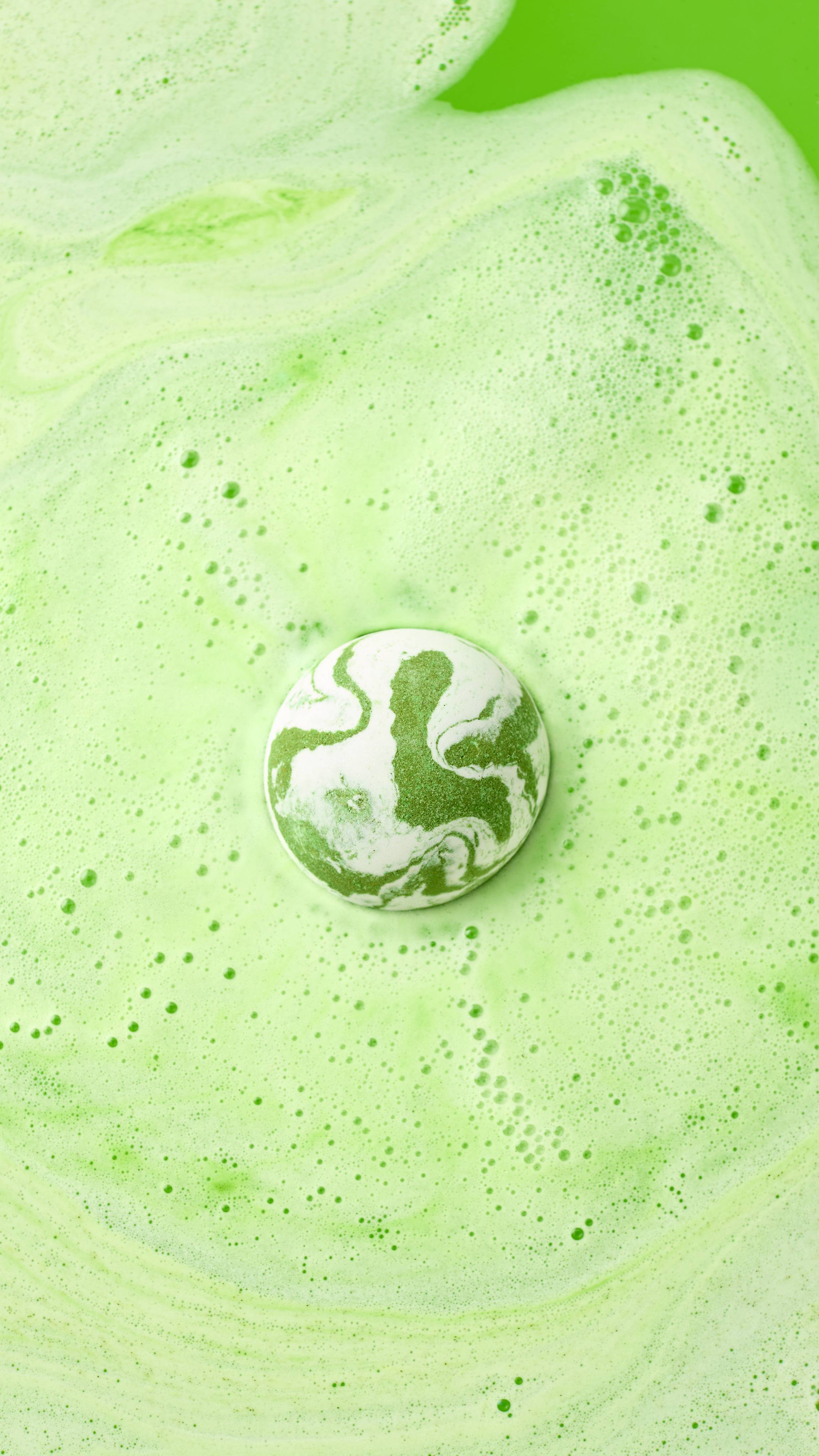 A hand holds the green and white marbled Cold Bath Bomb over a bathtub filled with vibrant green water. The bathwater swirls with light green foam, perfectly matching the colors of the bath bomb.