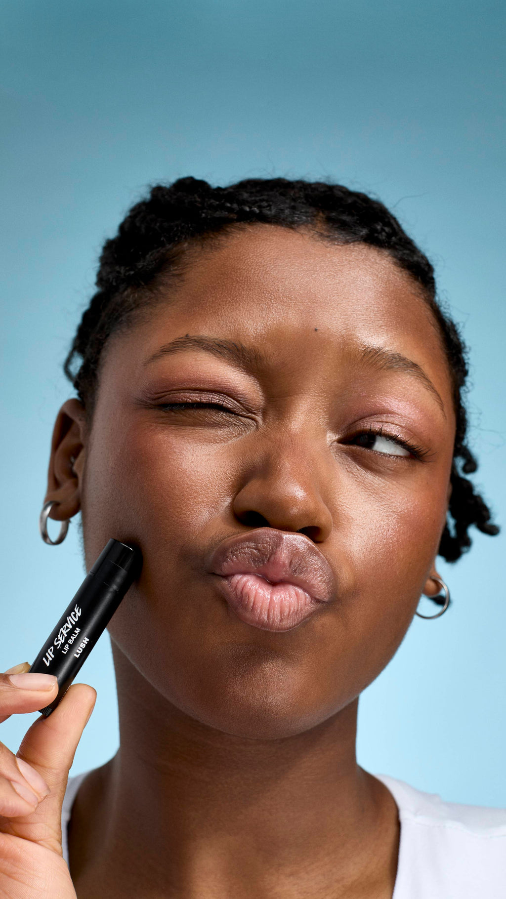 A close-up of a model with braided hair and glowing skin, making a playful pouting face with one eye shut. They hold a black tube of Lush 'Lip Service' lip balm against their cheek, set against a soft, light-blue background.