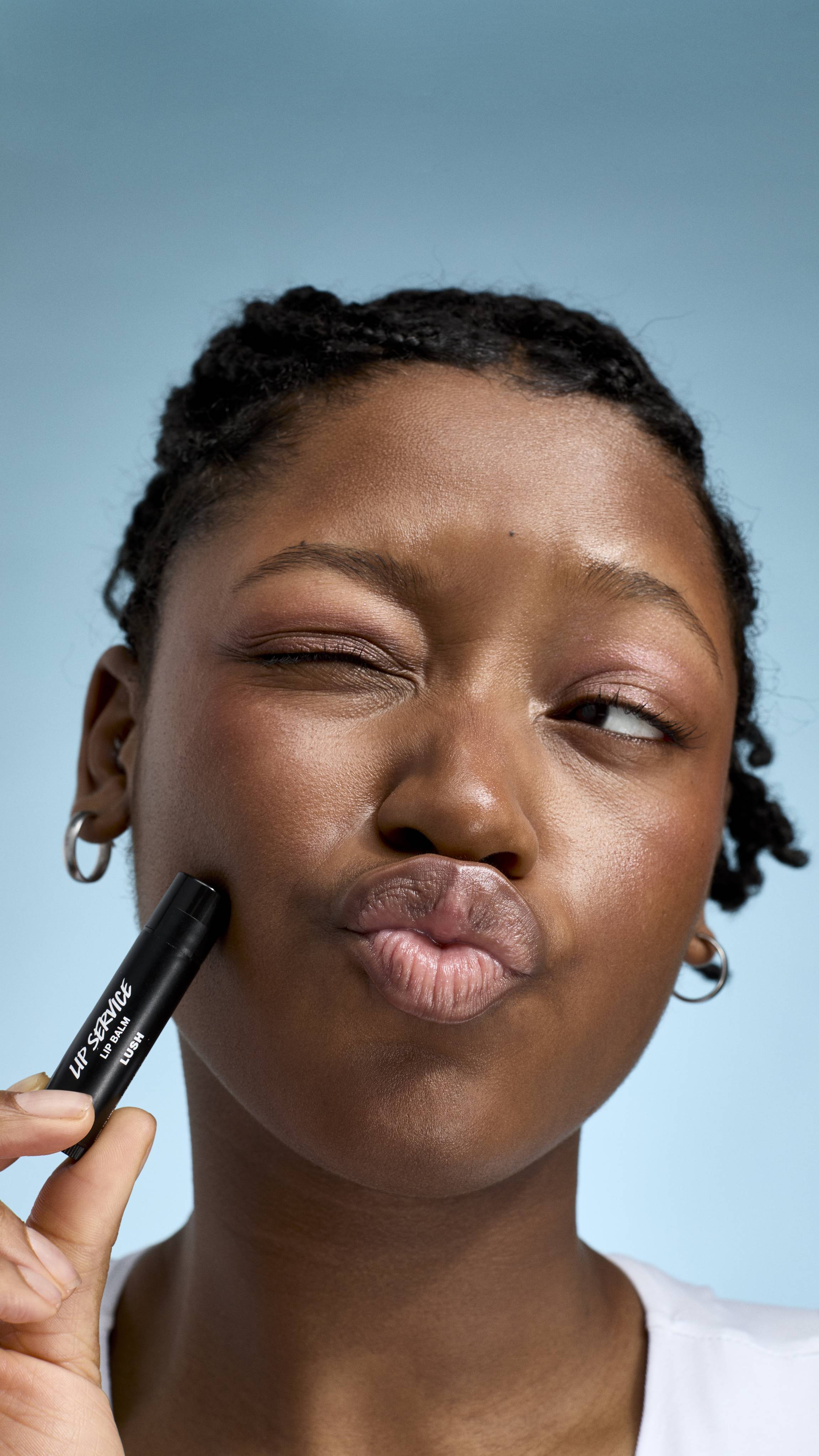 A close-up of a model with braided hair and glowing skin, making a playful pouting face with one eye shut. They hold a black tube of Lush 'Lip Service' lip balm against their cheek, set against a soft, light-blue background.