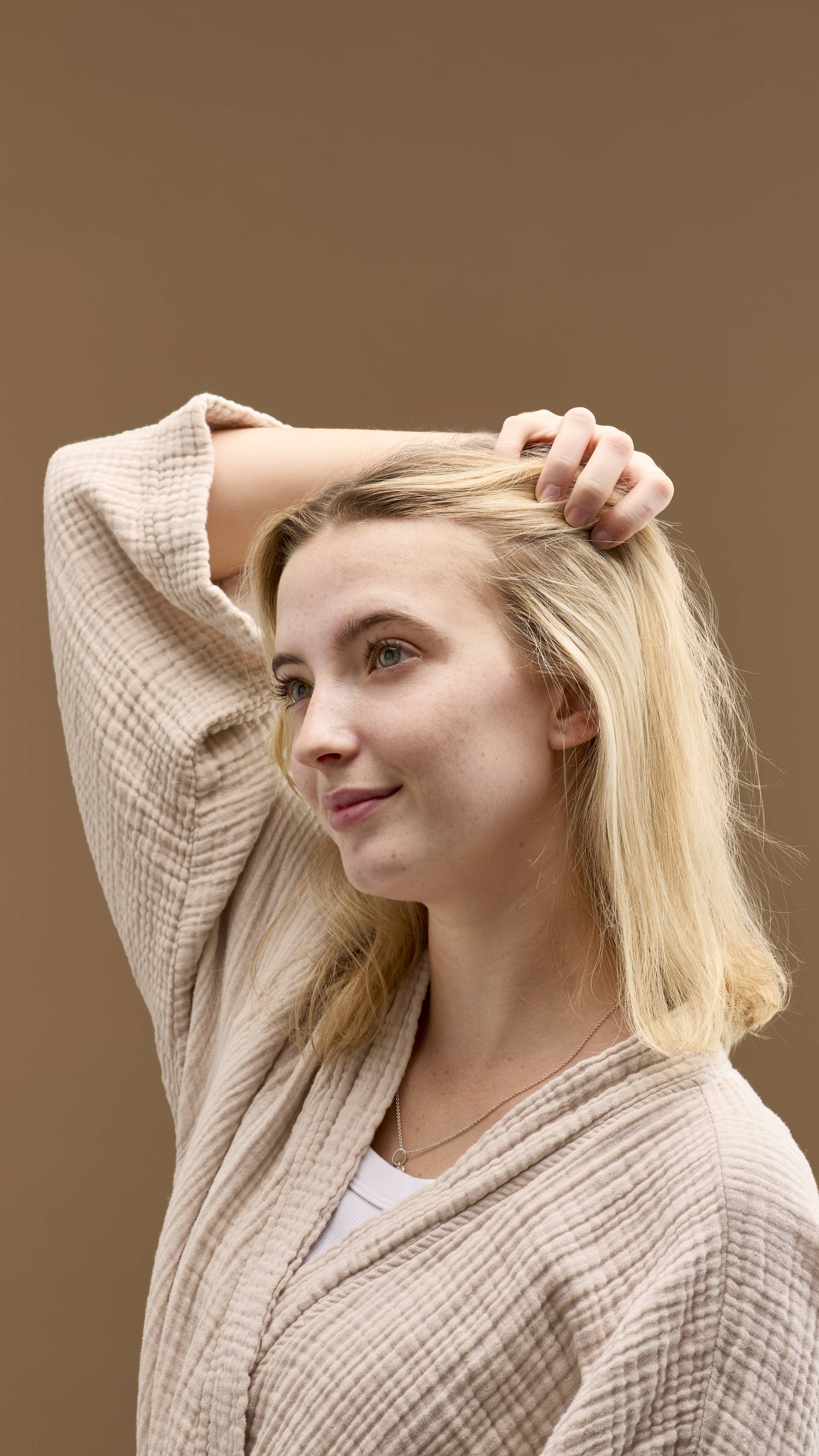 The image shows a person with light blonde hair, smiling. They run a hand through their hair, which appears clean and refreshed from the No Drought dry shampoo. They wear a light-coloured robe, against a solid beige background.