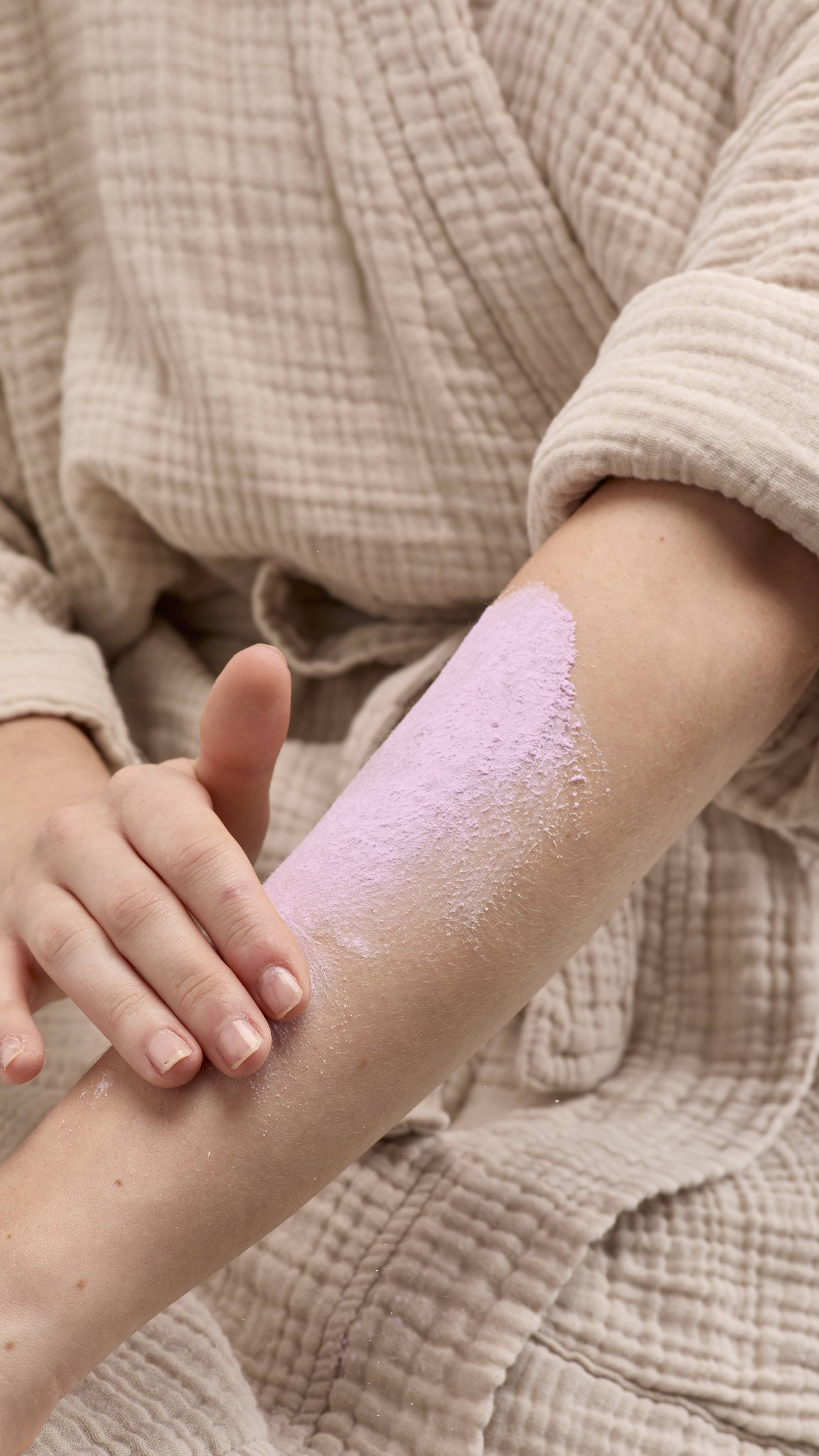 The image shows a hand rubbing a broad smear of pale purple 'Sleepy Dust' dusting powder onto a bare forearm. The person wears a light-coloured robe against a beige background.