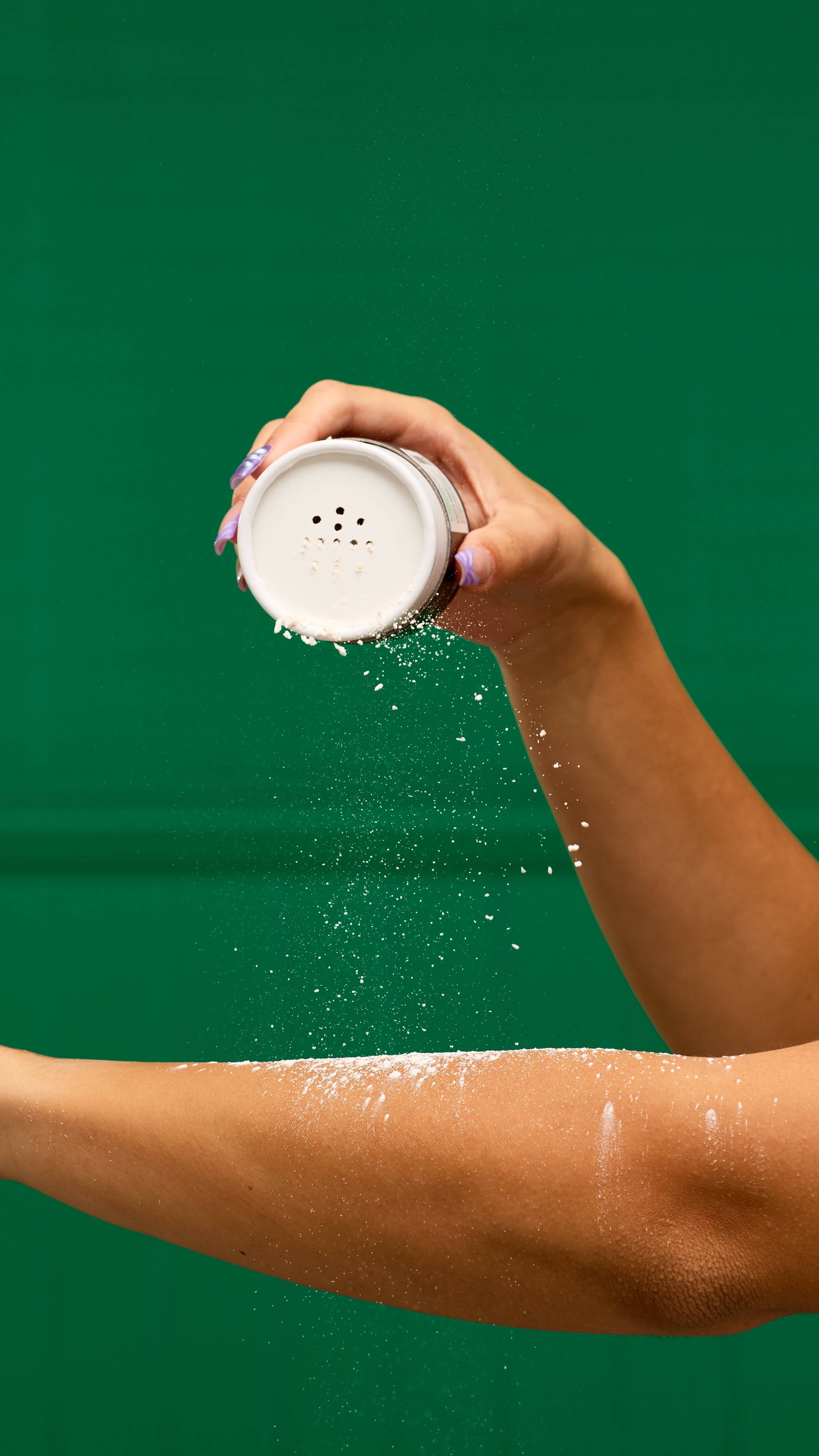 A person's hand shakes a LUSH Sticky Dates dusting powder container, with a fine cloud of white powder falling onto their forearm. The background is a vibrant green.