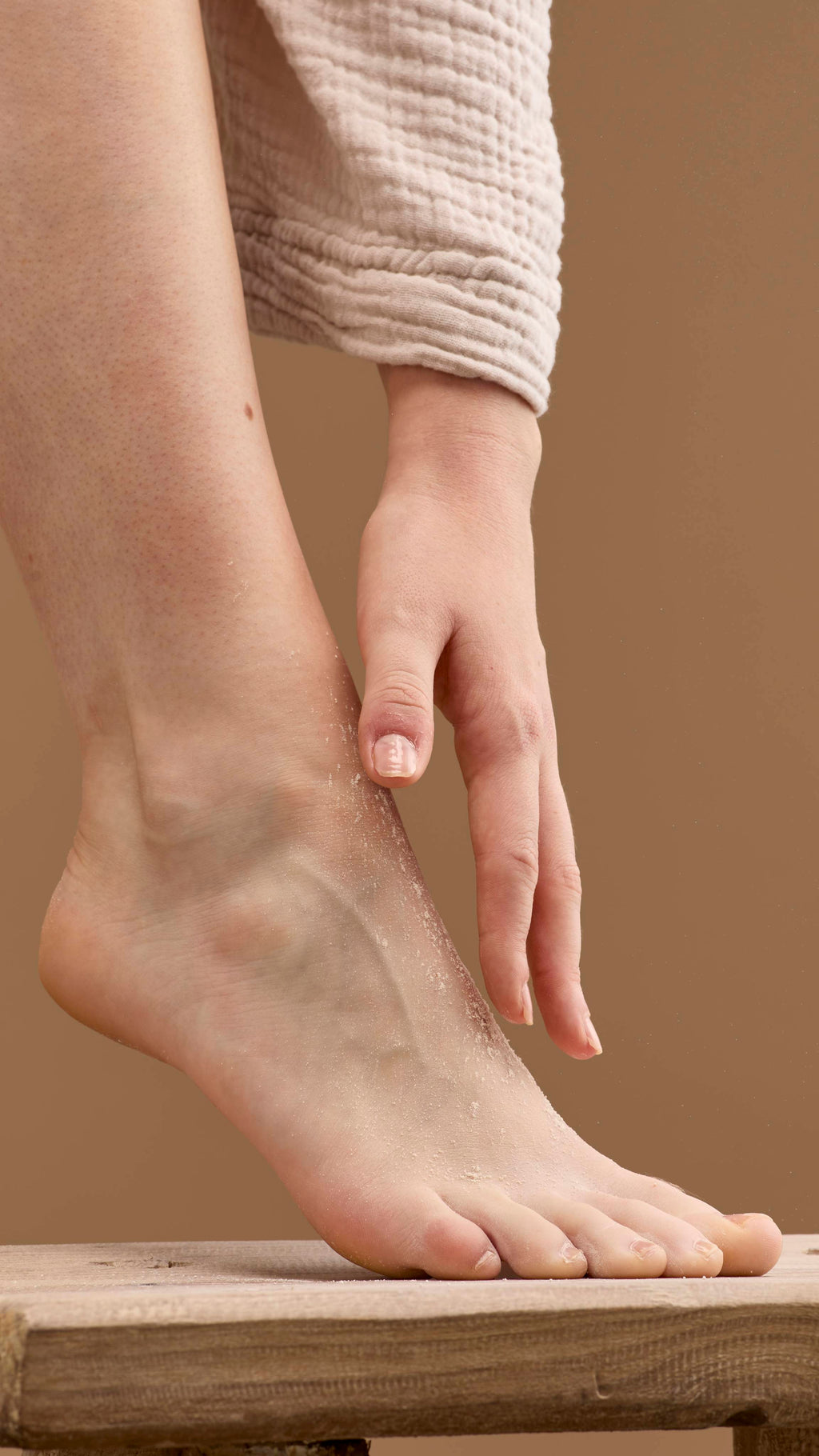 The image shows a hand rubbing a pale cream 'T For Toes' foot powder onto a bare foot and ankle. The foot rests on a wooden stool, with the person's robe visible against a solid beige background.