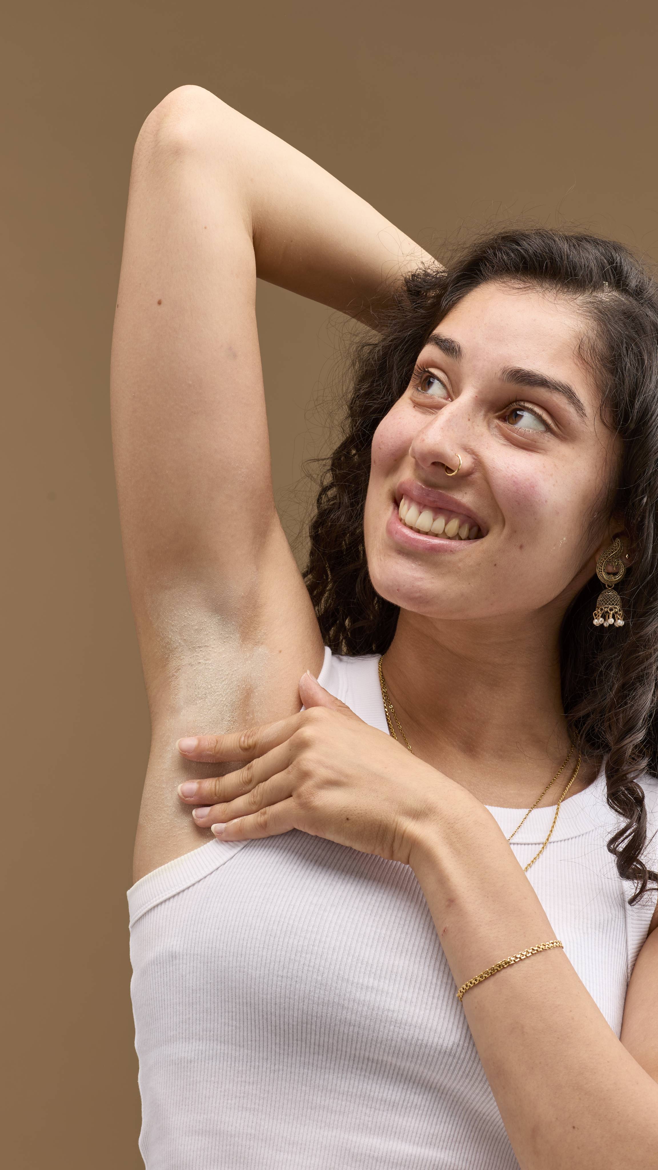 The image shows a person with dark, curly hair, smiling. They rub pale cream 'The Greeench' deodorant powder onto their raised underarm. The person wears a white vest top and necklaces against a light beige background.