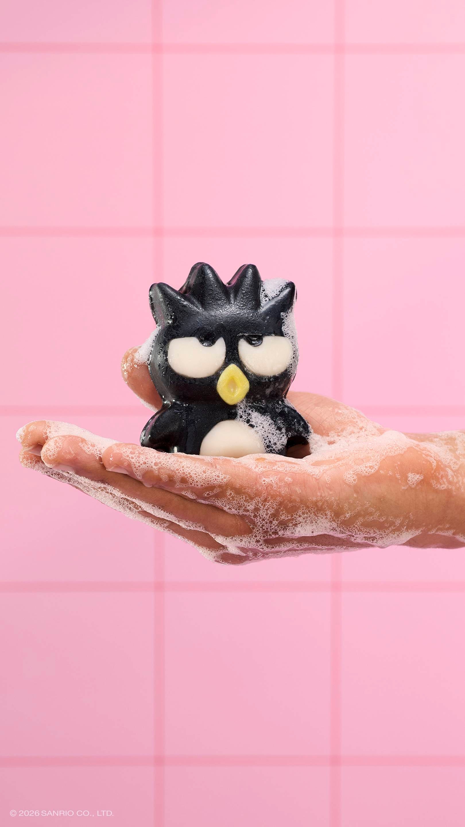 A soapy hand holds the black penguin-shaped "Bad Badtz-Maru" character soap, covered in white bubbles. The scene is set against a background of pink square tiles.