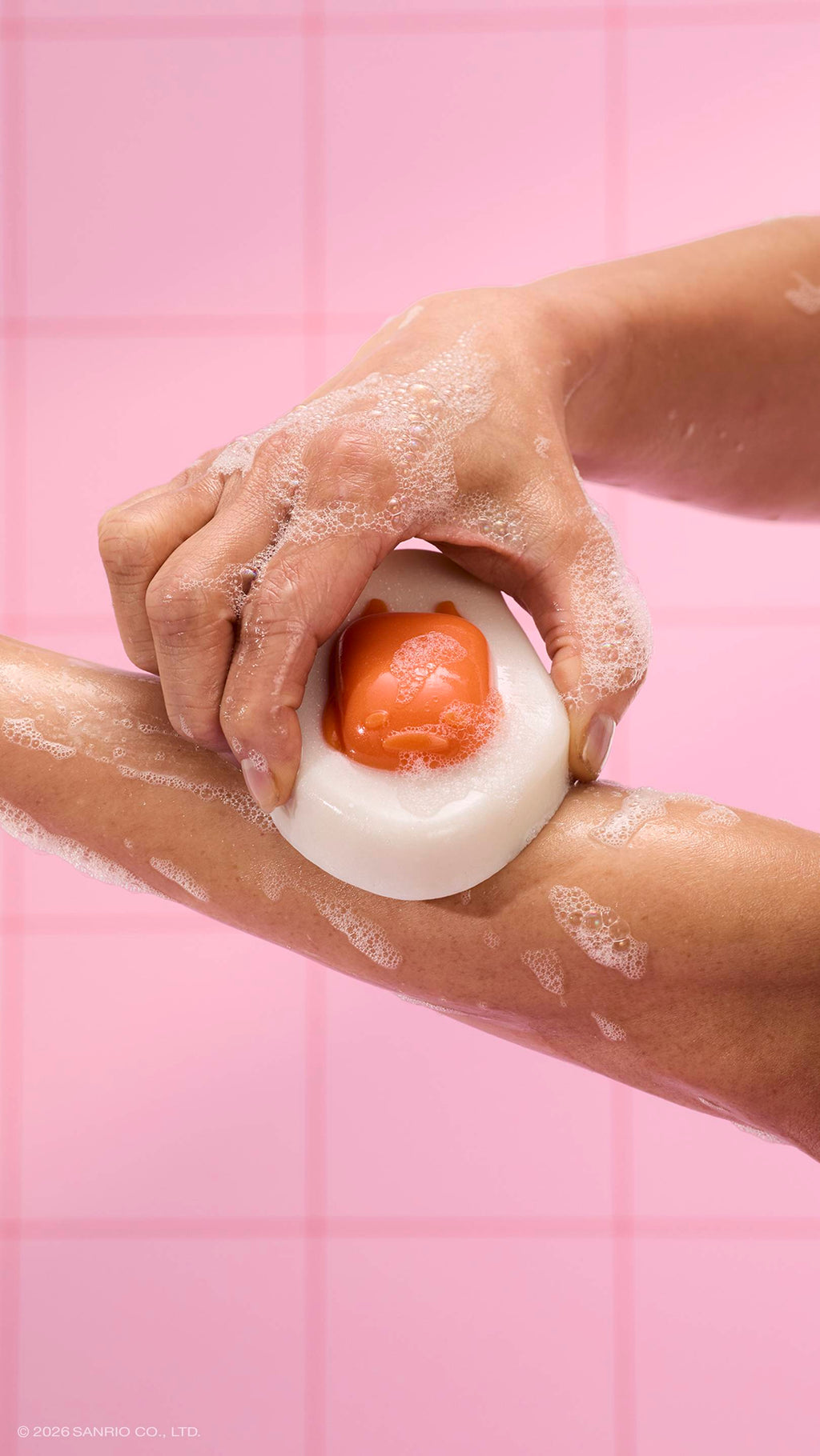 A close-up shows a person rubbing an egg-shaped "Gudetama" shower jelly against their arm. The glossy jelly features a sleepy orange face as the "yolk" and is covered in white bubbles, set against a pink tiled wall.