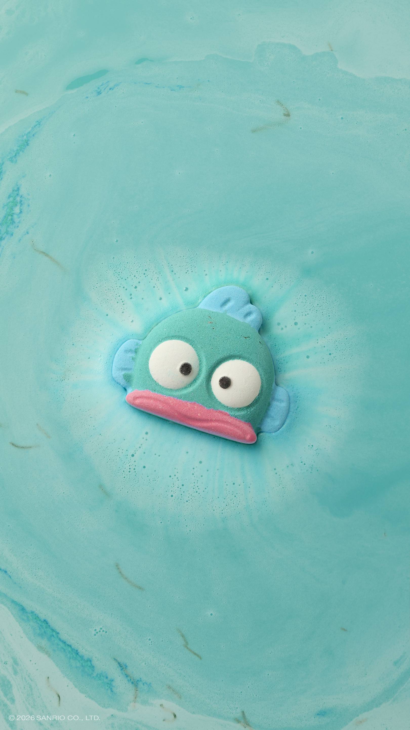 Hangyodon. A teal, fish-shaped bath bomb floats in a bath of turquoise water. It has large white eyes and a pink mouth, and is surrounded by a ring of white foam and bubbles as it dissolves.