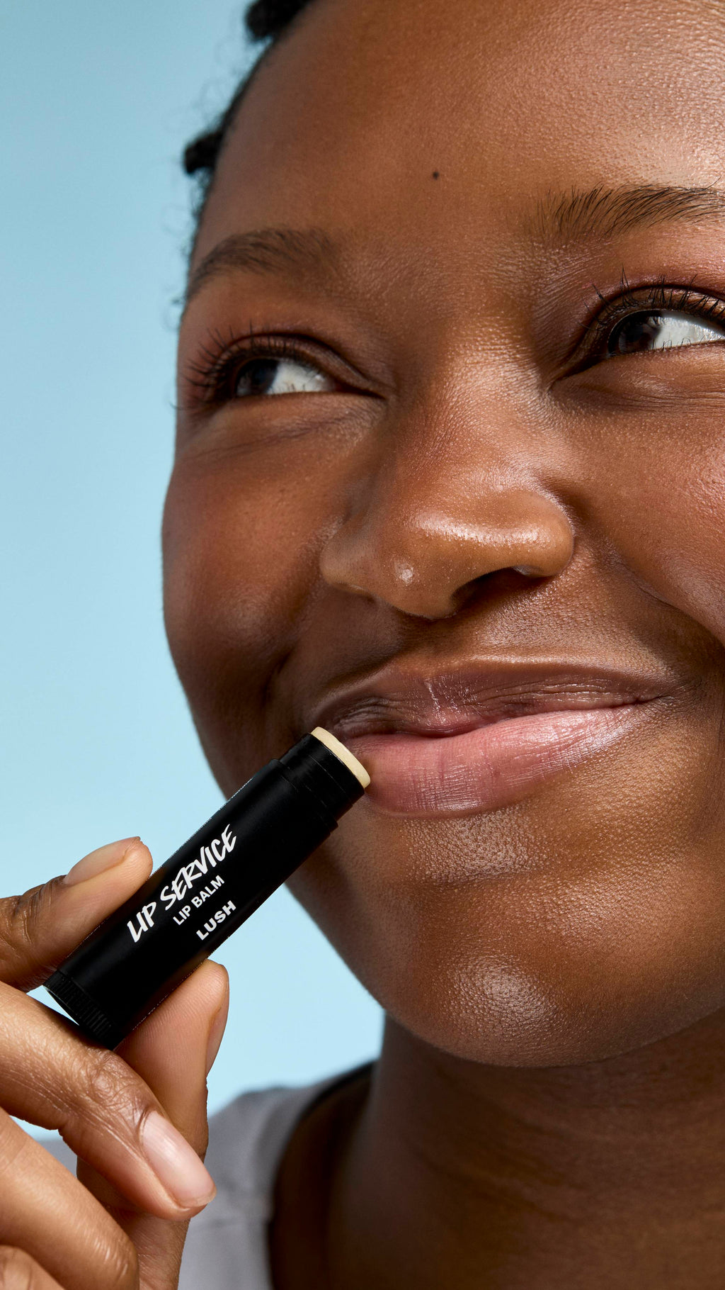 A close-up of a model applying 'Lip Service' lip balm to their lips. They have a soft smile and are looking slightly off-camera, holding the black Lush tube against their mouth against a clear, light-blue background.