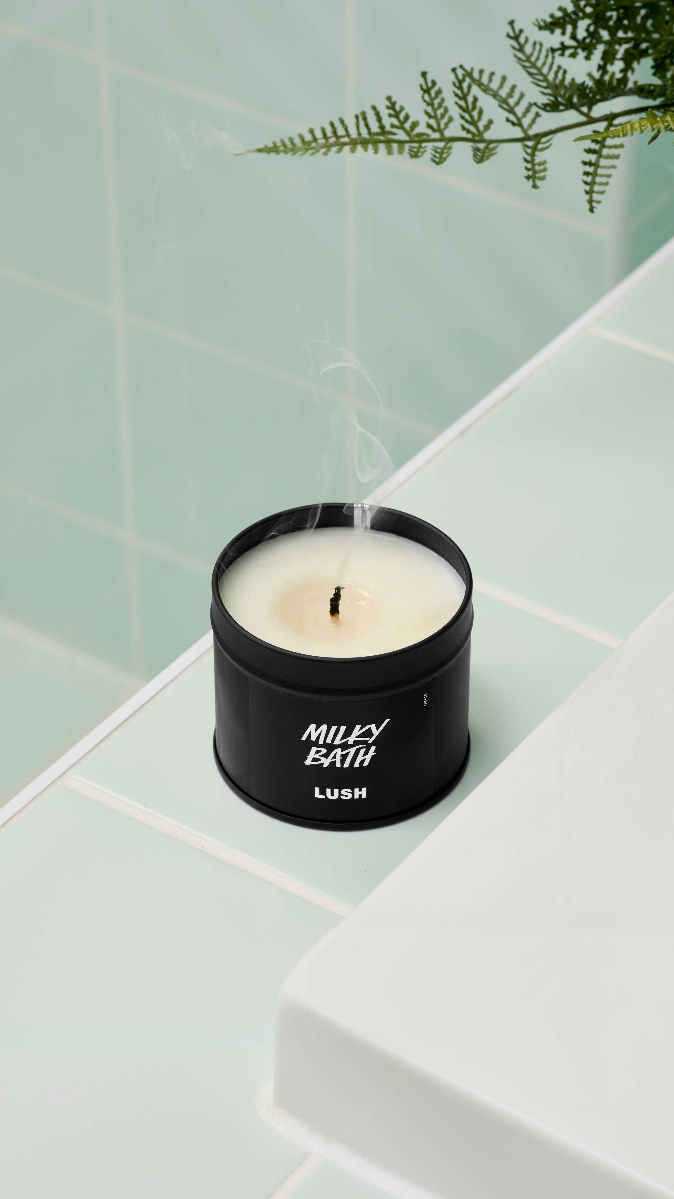 A close-up, high-angle shot of an extinguished Milky Bath candle by Lush in a matte black tin. Thin wisps of smoke rise from the blackened wick. It sits on a white ledge against mint green wall tiles with a fern leaf visible at the top.