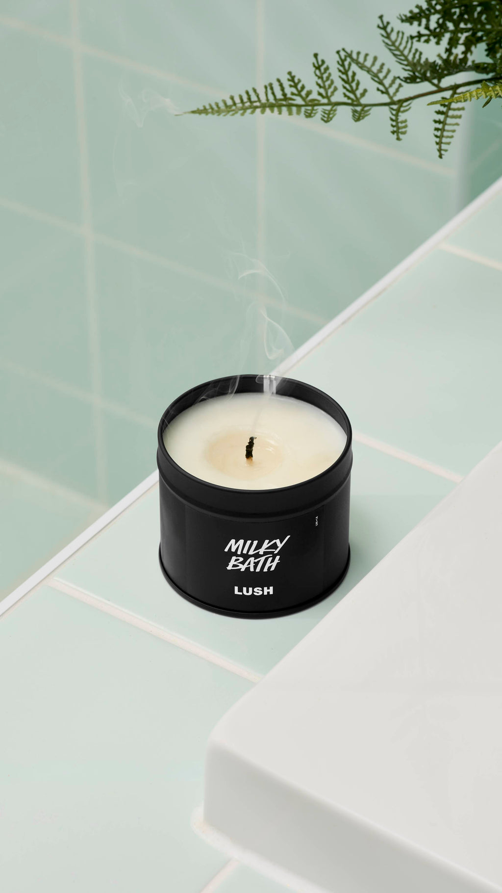 A close-up, high-angle shot of an extinguished Milky Bath candle by Lush in a matte black tin. Thin wisps of smoke rise from the blackened wick. It sits on a white ledge against mint green wall tiles with a fern leaf visible at the top.