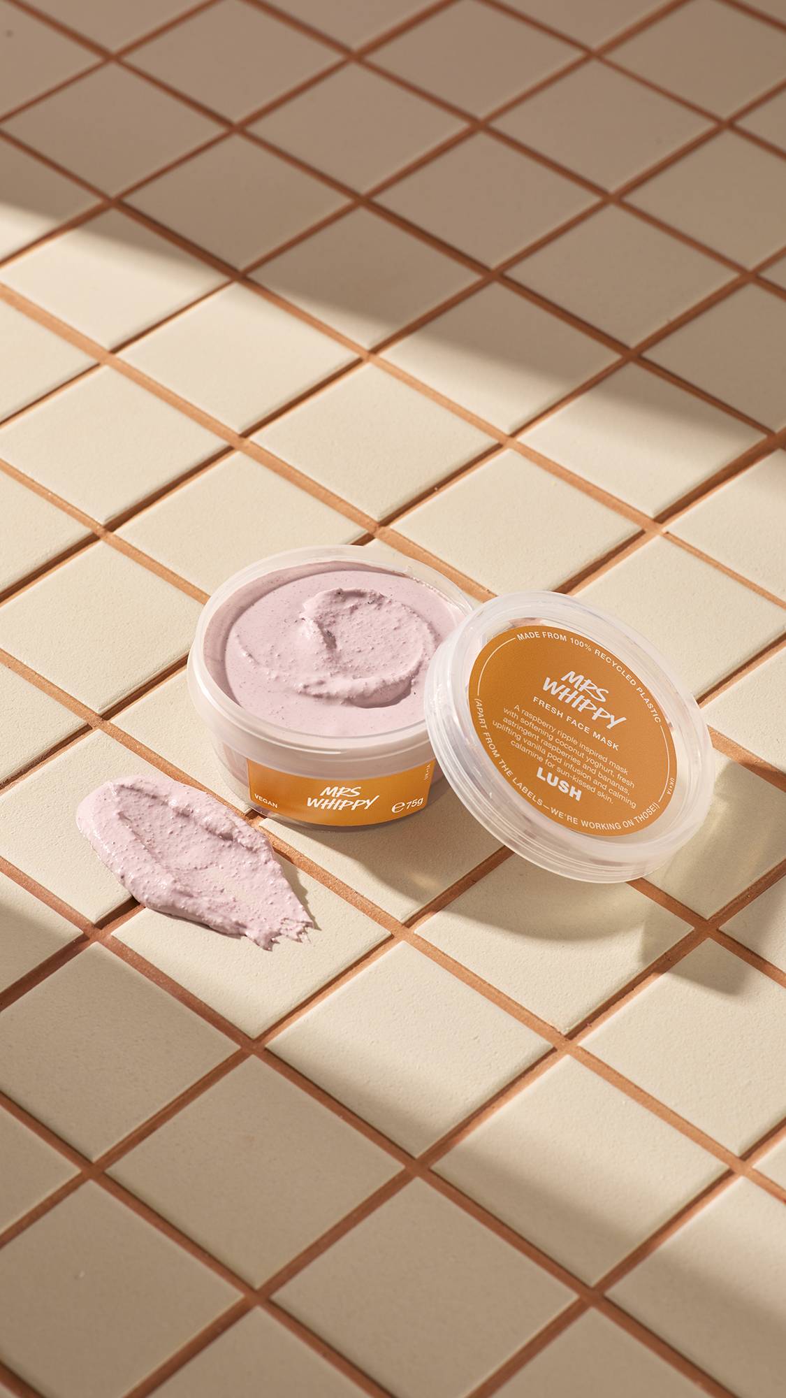 The image shows a clear, recycled plastic, open pot of Mrs Whippy face mask with orange labels, the lid leaning on the side and a smeared swatch of the textured product on the cream-tiled floor beside the pot.