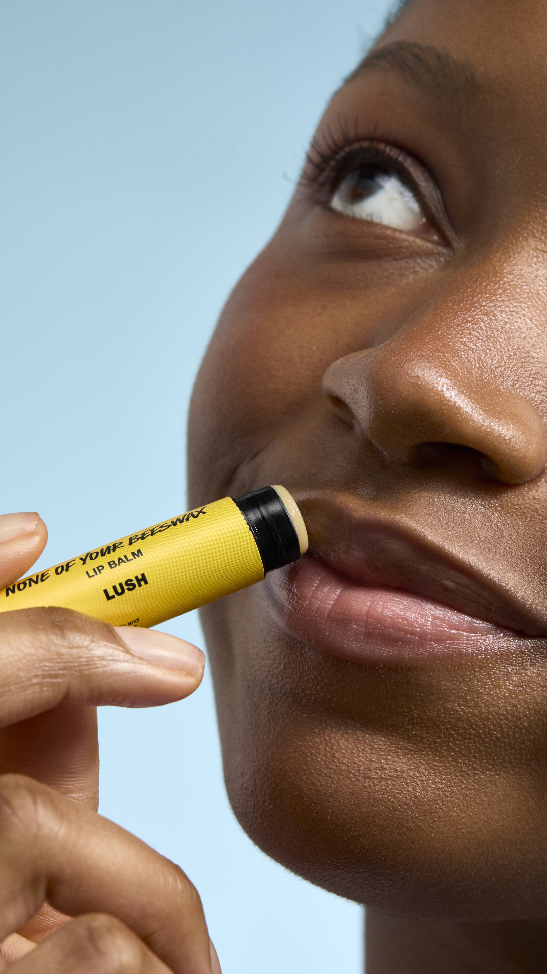 A close-up of a model applying 'None of Your Beeswax' lip balm to their lips. They are looking upwards with a soft smile, holding the bright yellow Lush tube against their mouth. The background is a clear, light blue.