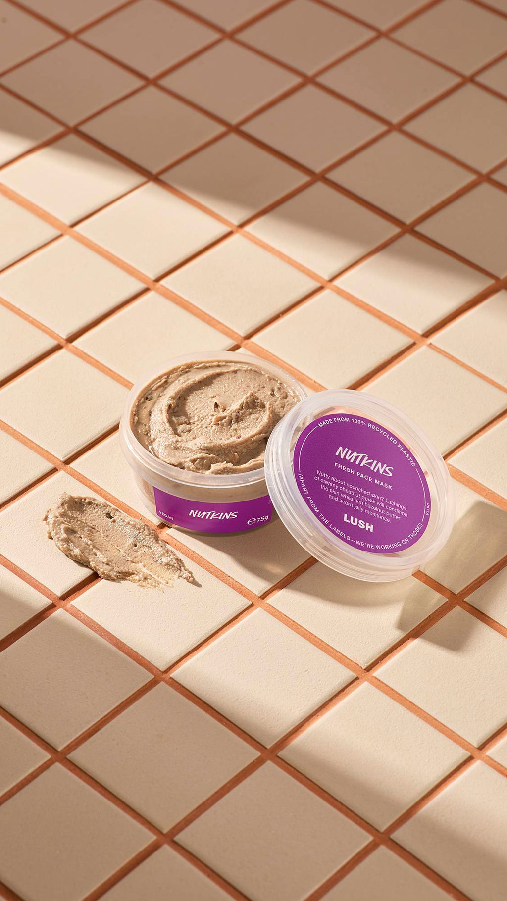 The image shows a clear, recycled plastic, open pot of Nutkins face mask with purple labels, the lid leaning on the side and a smeared swatch of the textured product on the cream-tiled floor beside the pot.