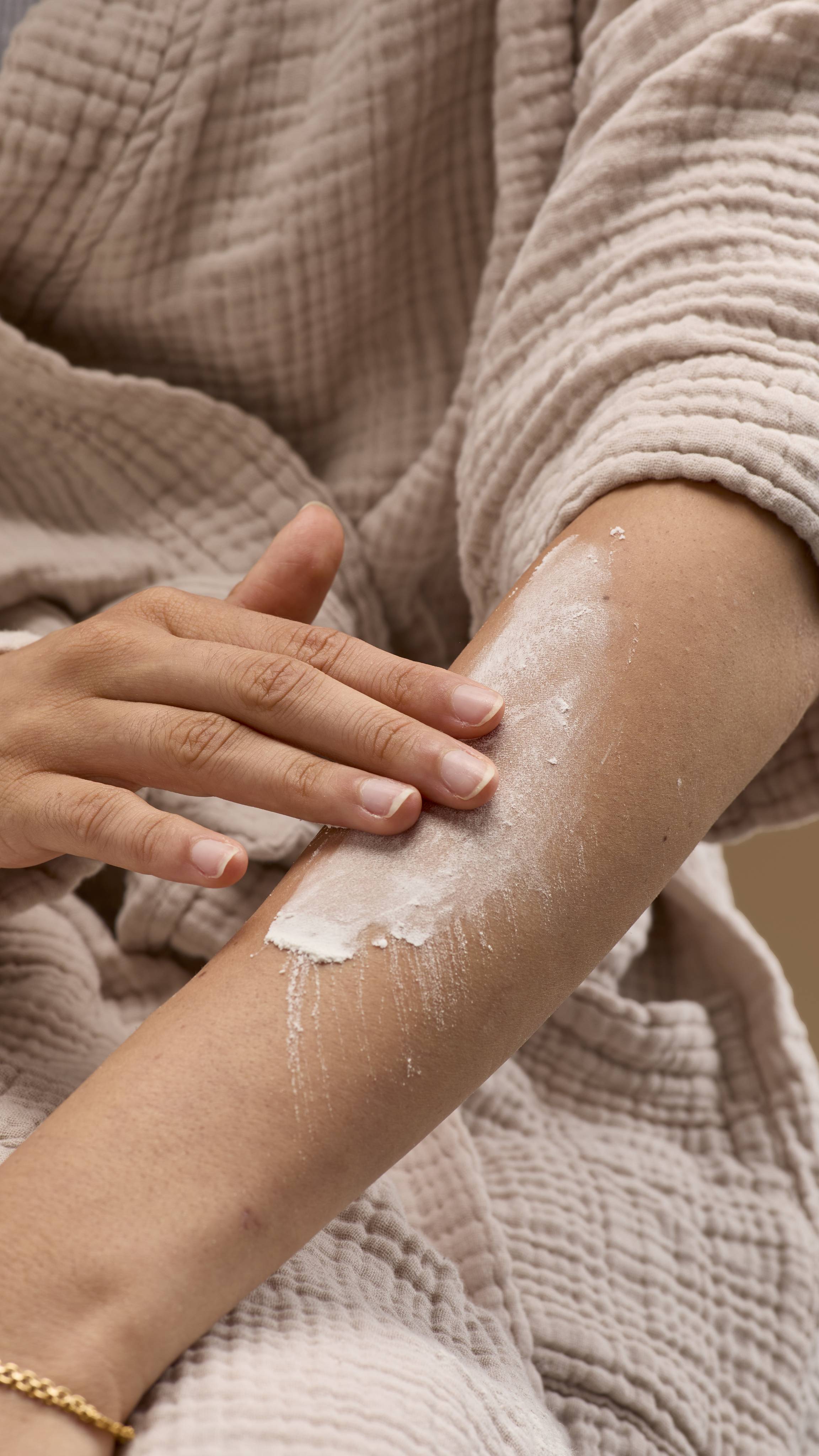 The image shows a hand rubbing a broad smear of pale cream 'Silky Underwear' dusting powder onto a bare forearm. The person wears a light-coloured robe and bracelet against a solid beige background.