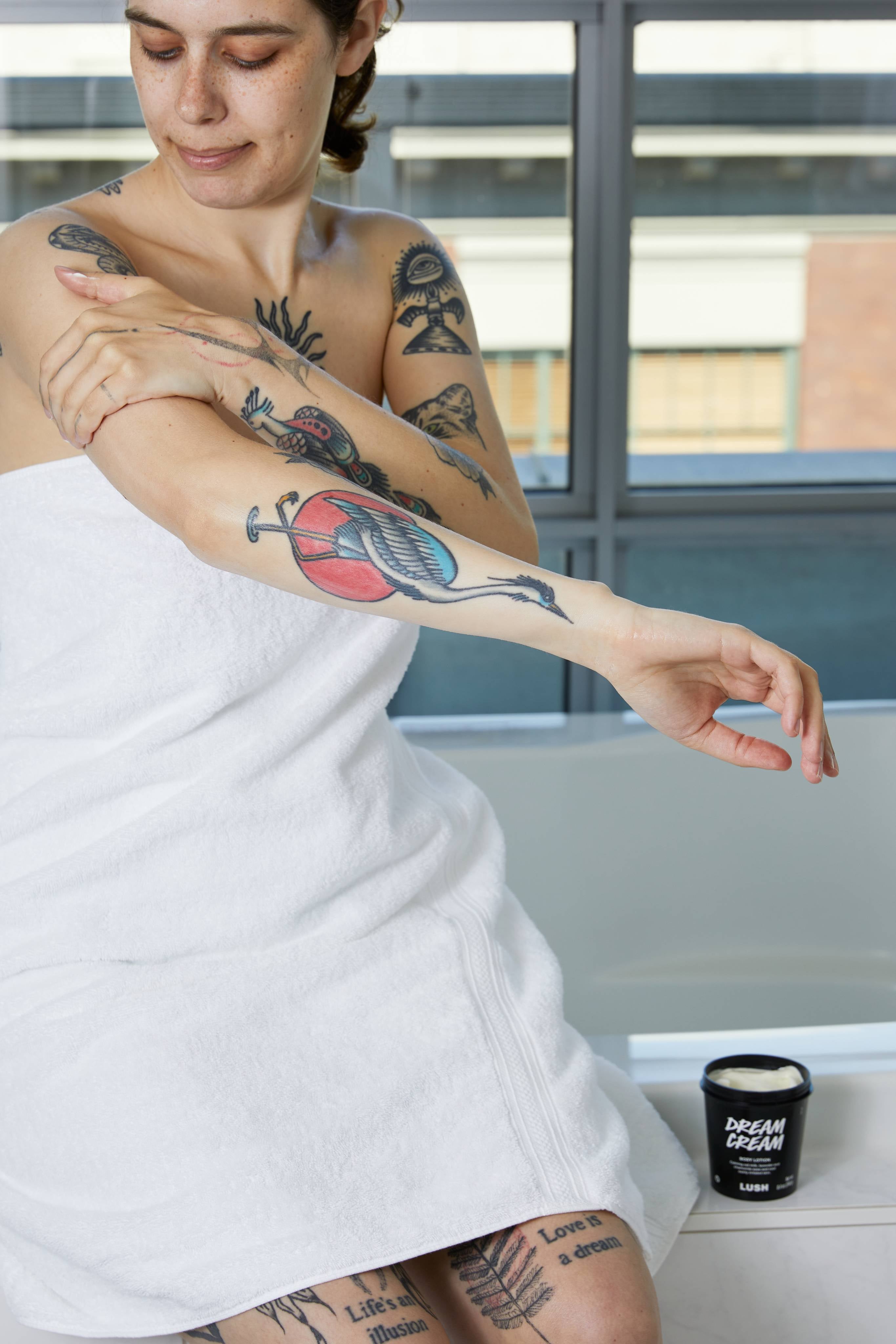 A tattooed person wrapped in a white towel sits on the edge of a bath, massaging cream into their arm. An open black tub of Lush 'Dream Cream' body lotion sits beside them on the tiled ledge.