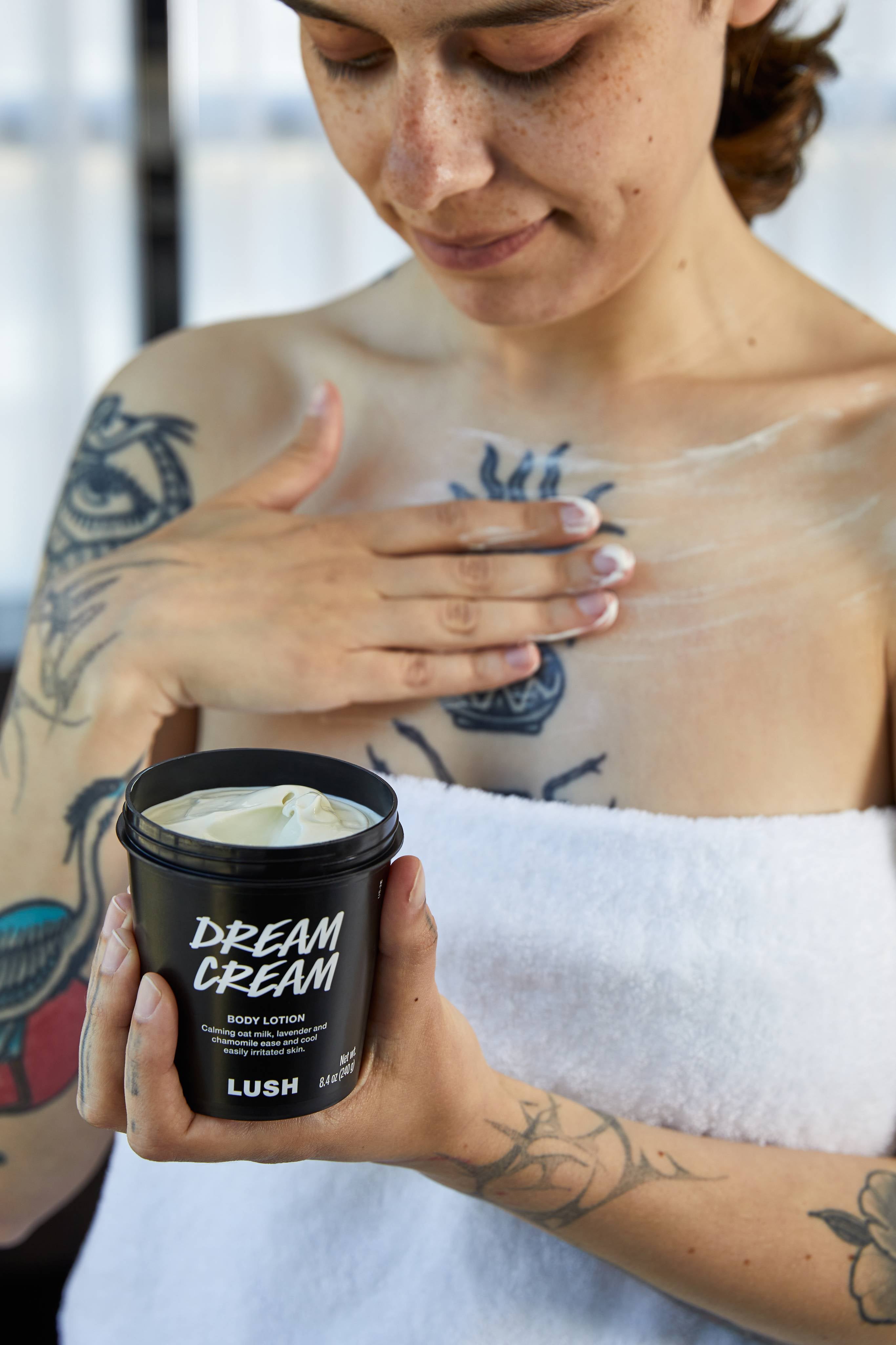 A tattooed person wrapped in a white towel smooths pale lotion onto their chest. With their other hand, they hold an open black tub of Lush 'Dream Cream' body lotion, looking down with a gentle smile.