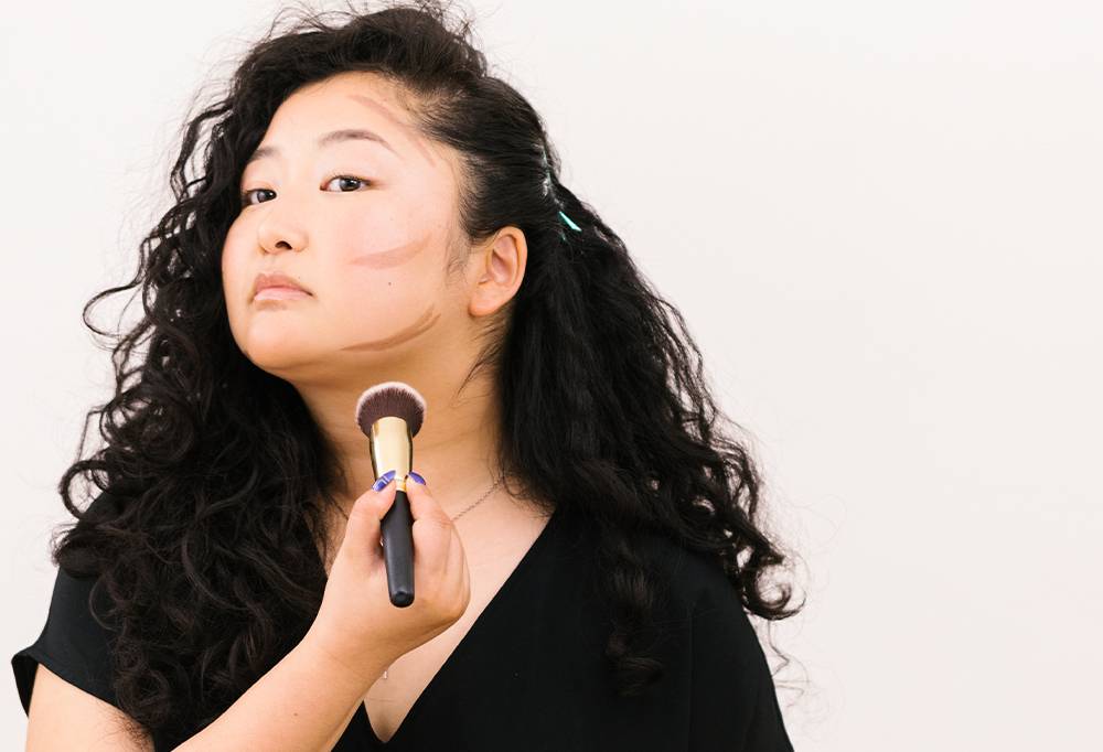 A person with long, dark hair holds a BFF Brush, ready to buff out the dark contours drawn onto their cheeks and jawline.