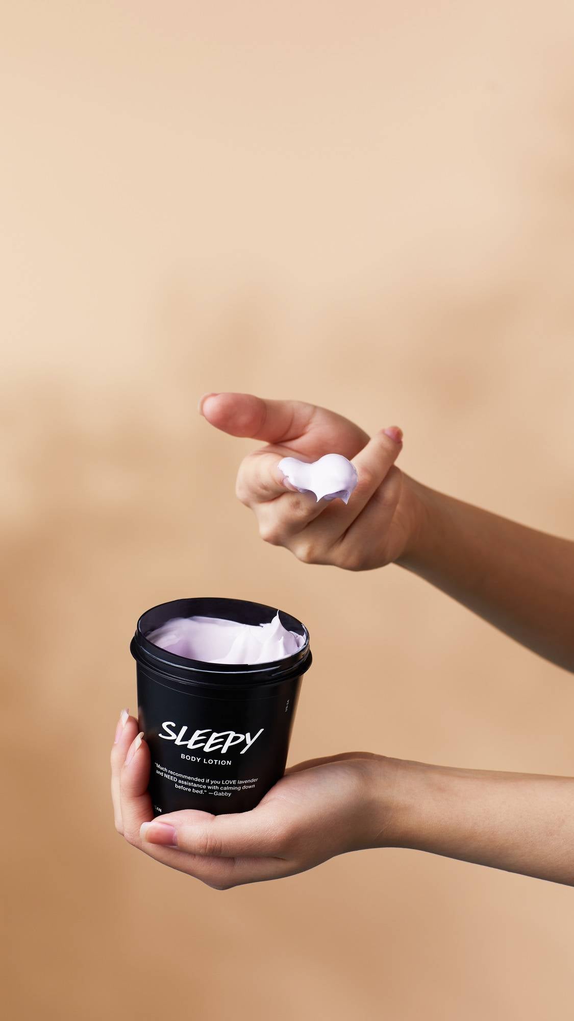 Hands hold an open black pot of Lush Sleepy body lotion against a warm beige background. One hand balances the tub, while the other holds up a dollop of pale purple cream scooped onto the fingers.